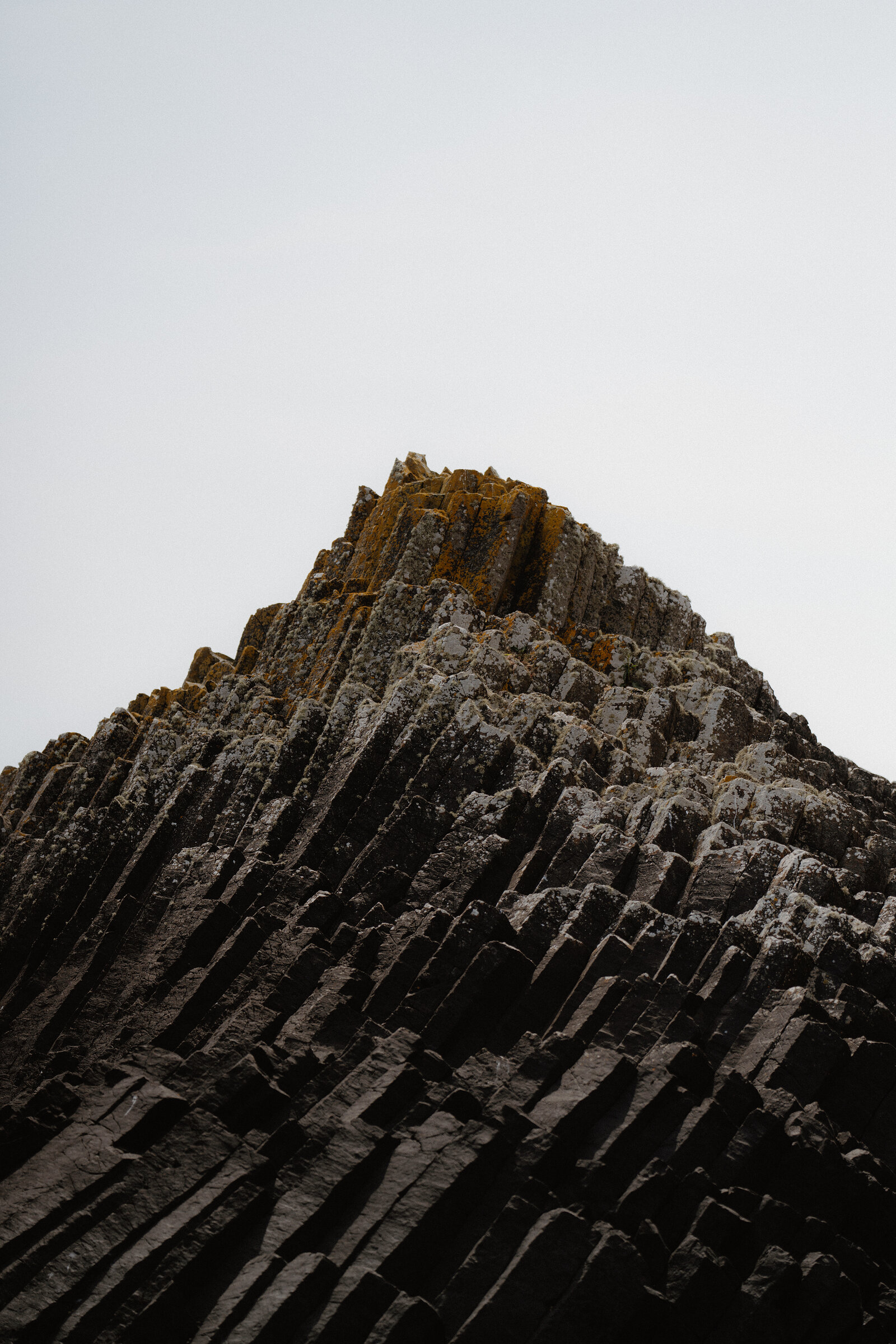 A jagged basalt pinnacle rises to a lichen-streaked point, its columns fanning out below against a pale sky.
