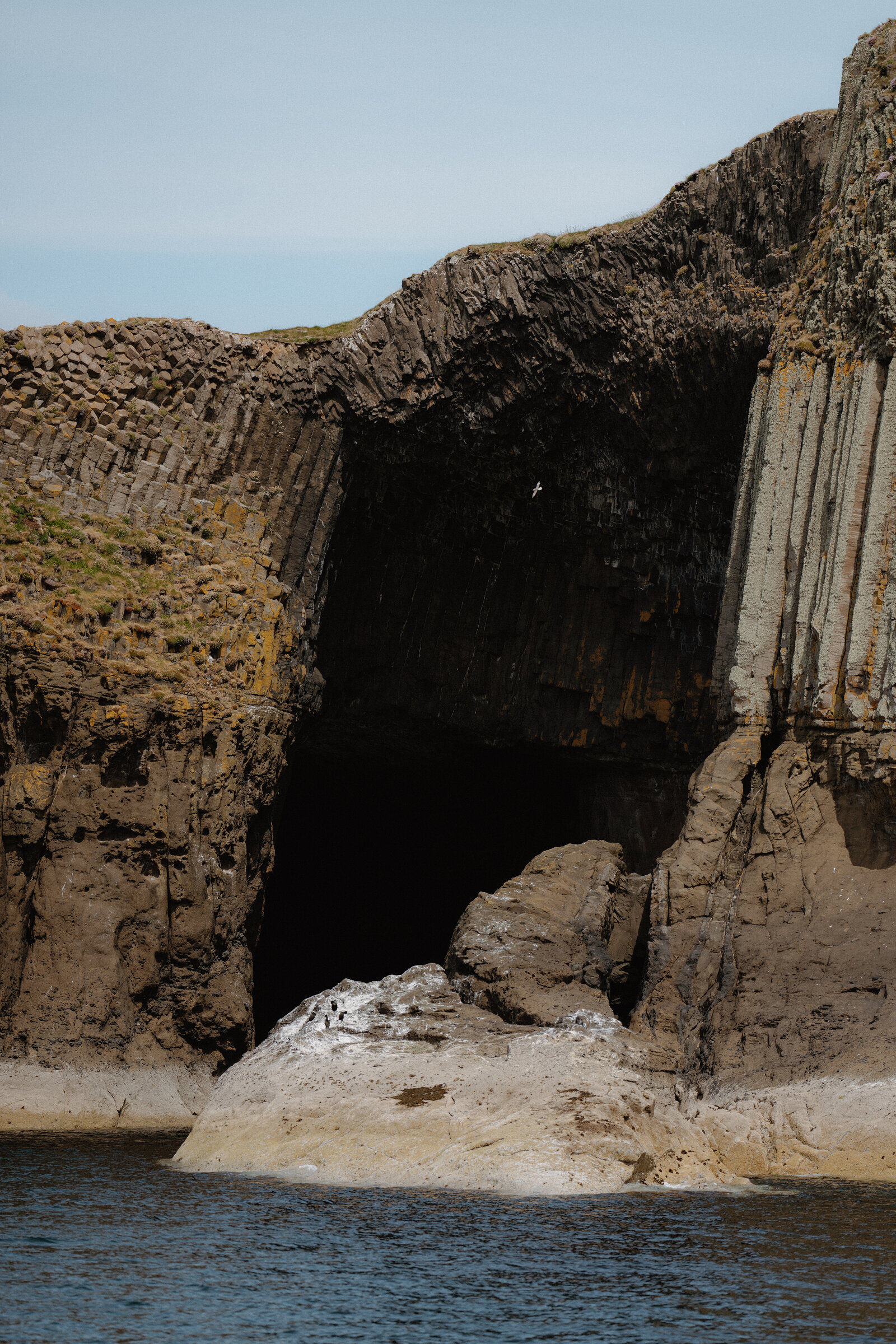 A dark sea cave mouth opens beneath tiered basalt columns, a pale rock spit reaching out from its entrance.