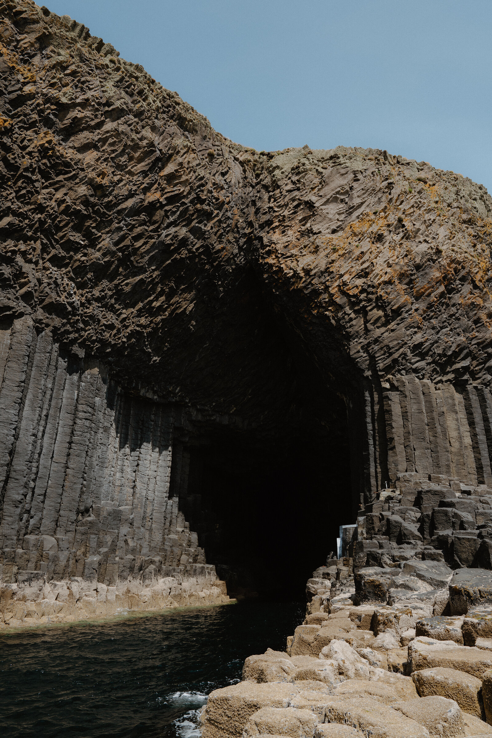 Hexagonal basalt columns form the mouth of a sea cave, dark water reaching into the shadowed interior beneath a rugged clifftop.