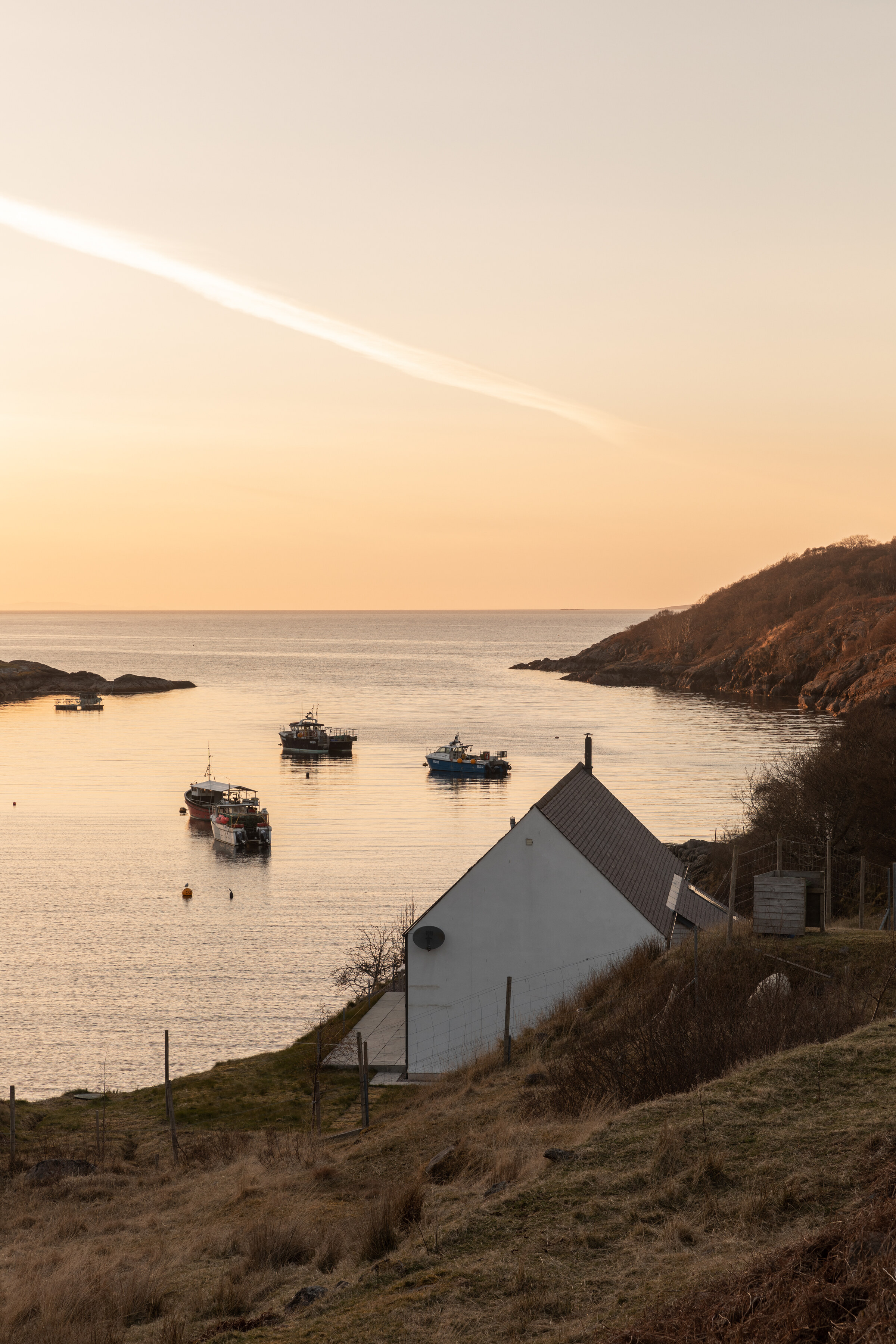 Fishing boats lie at their moorings off Ardheslaig, below The Net Store’s slate roof, in the evening light.