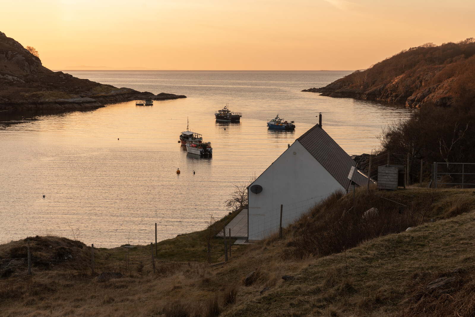 The Net Store’s white gable stands on the shore at Ardheslaig above Loch Beag, with fishing boats moored in the bay at dusk.