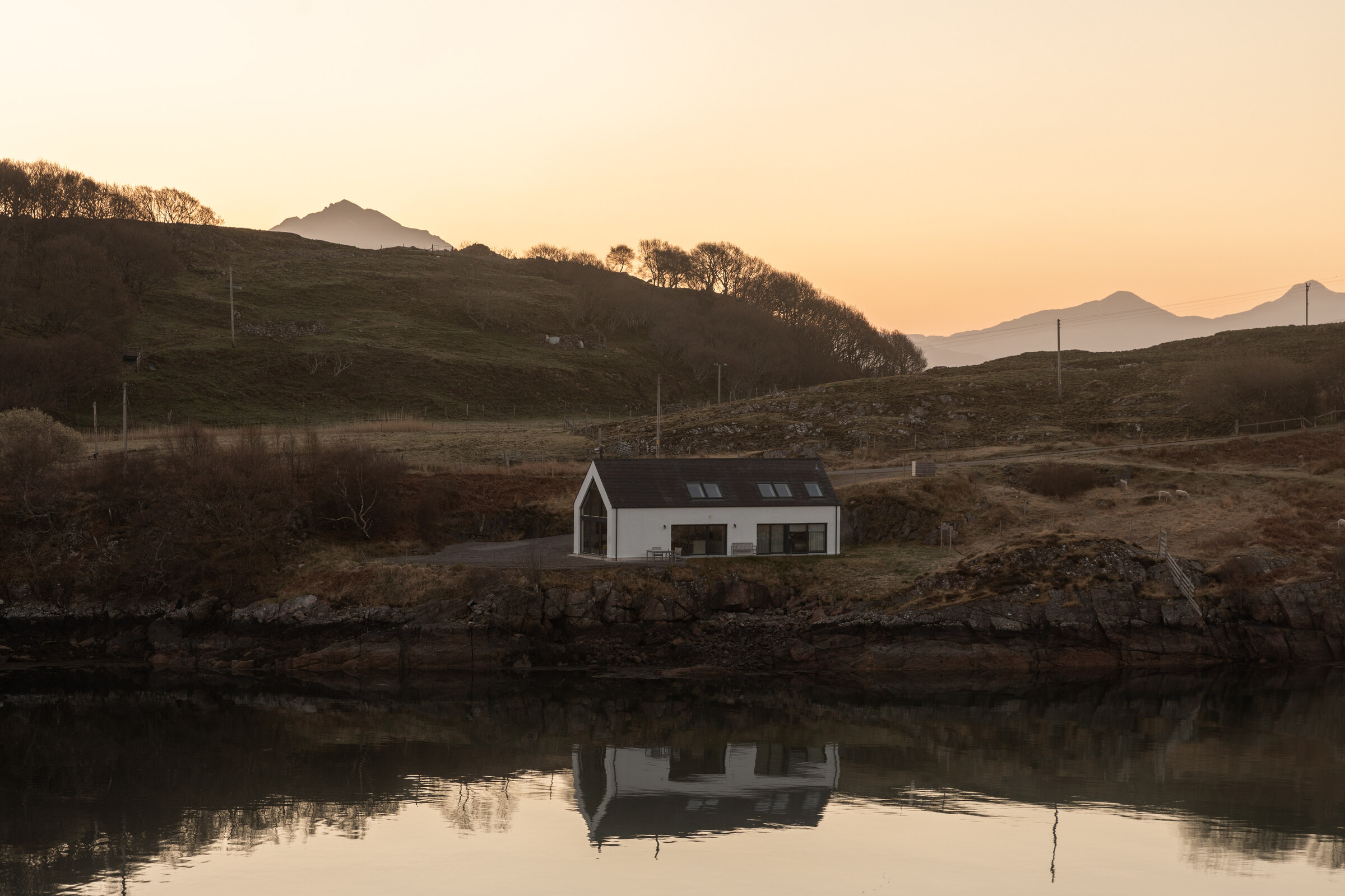 The Net Store, small and white on the far shore, reflected in the still water of Loch Beag, with hills behind.