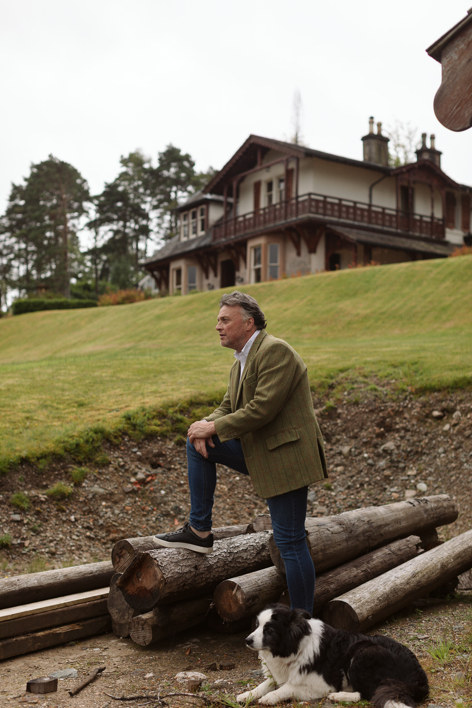 Iain Hopkins rests a foot on a pile of logs below the lodge at Woodside, his border collie lying at his feet.