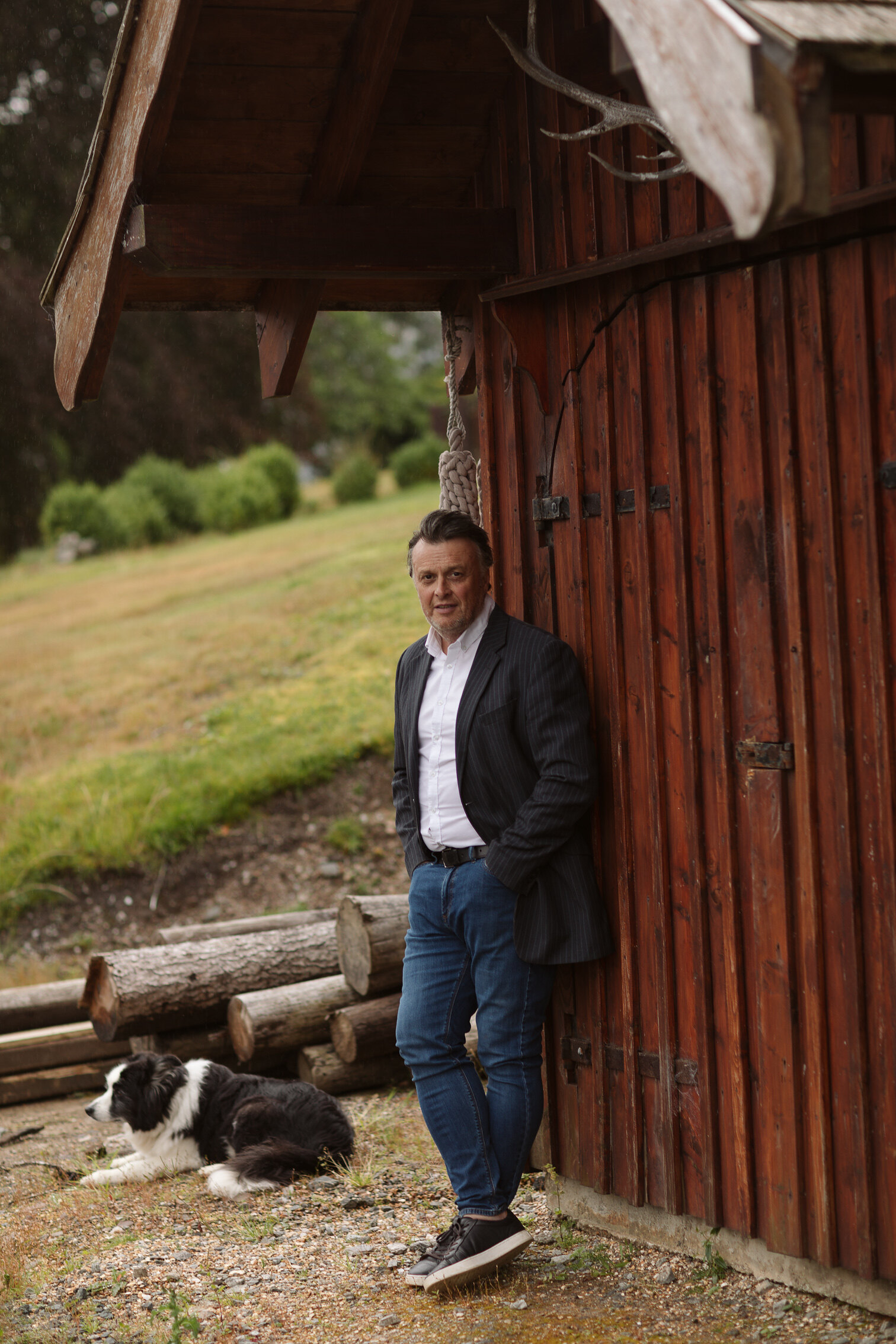 Iain Hopkins leans against the red timber boathouse at Woodside Lodge, his border collie lying by a pile of logs.