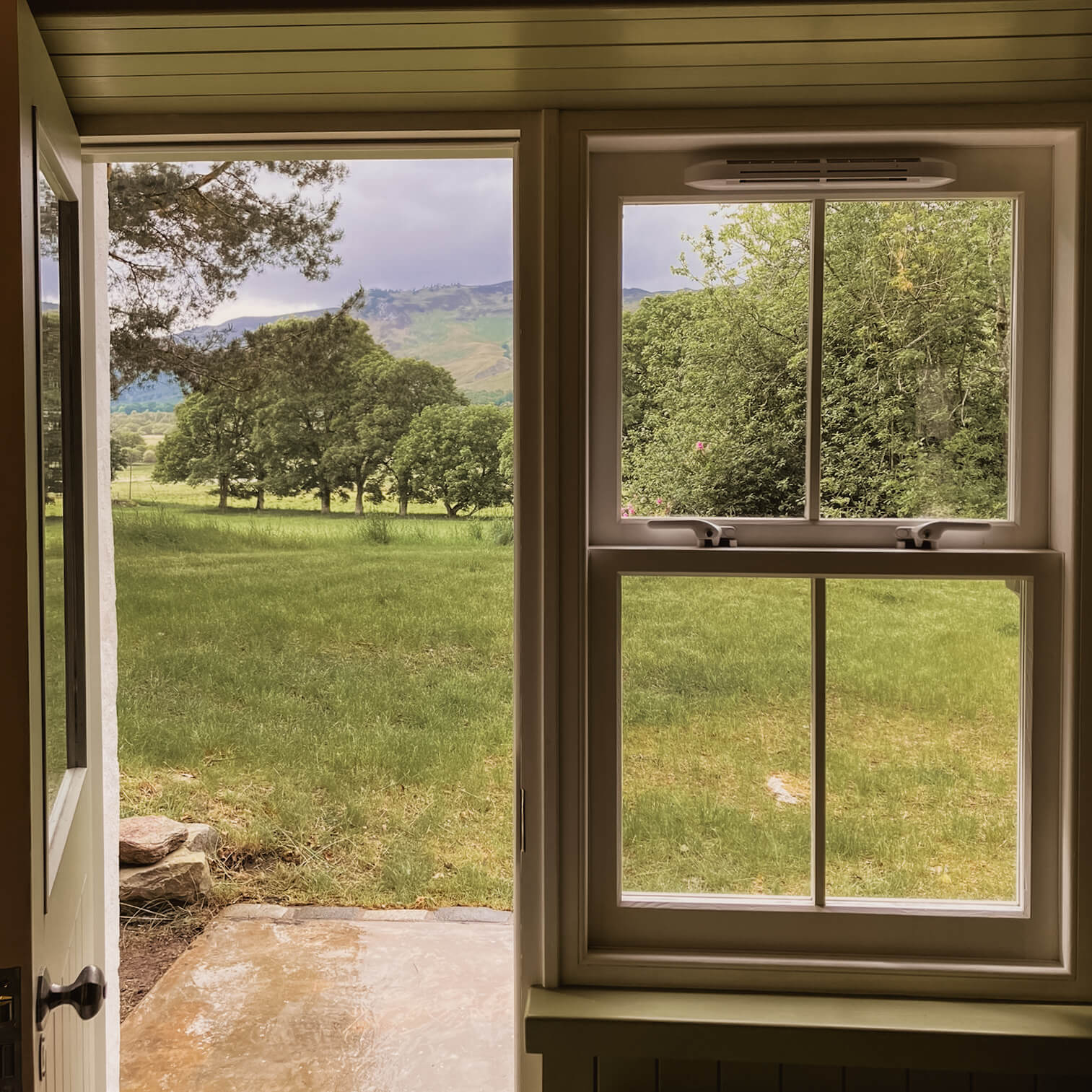 An open door and a sash window look out from the bothy across a field and trees to the hills.