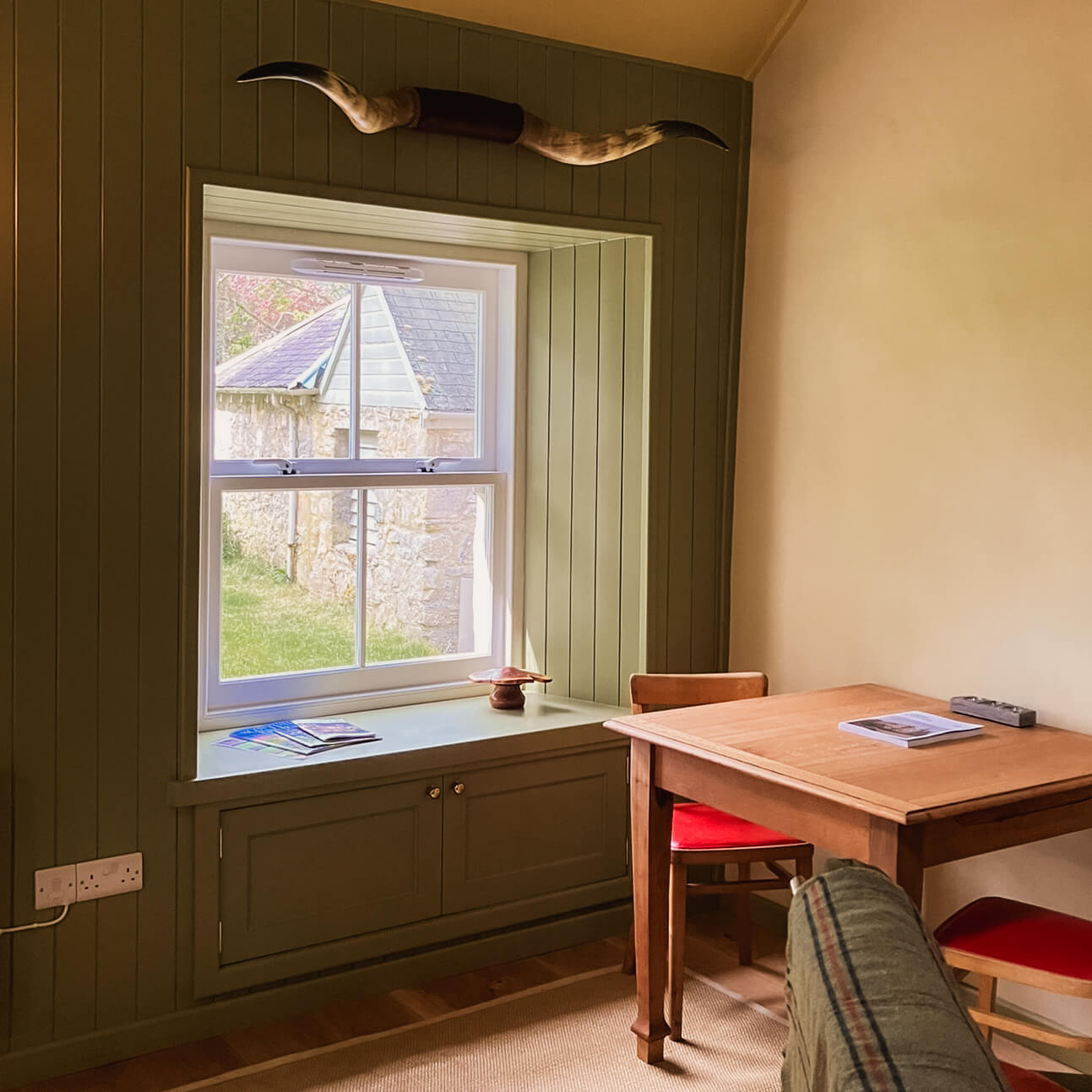A small table and red chairs stand by a sash window set in green panelling, a stone building outside.