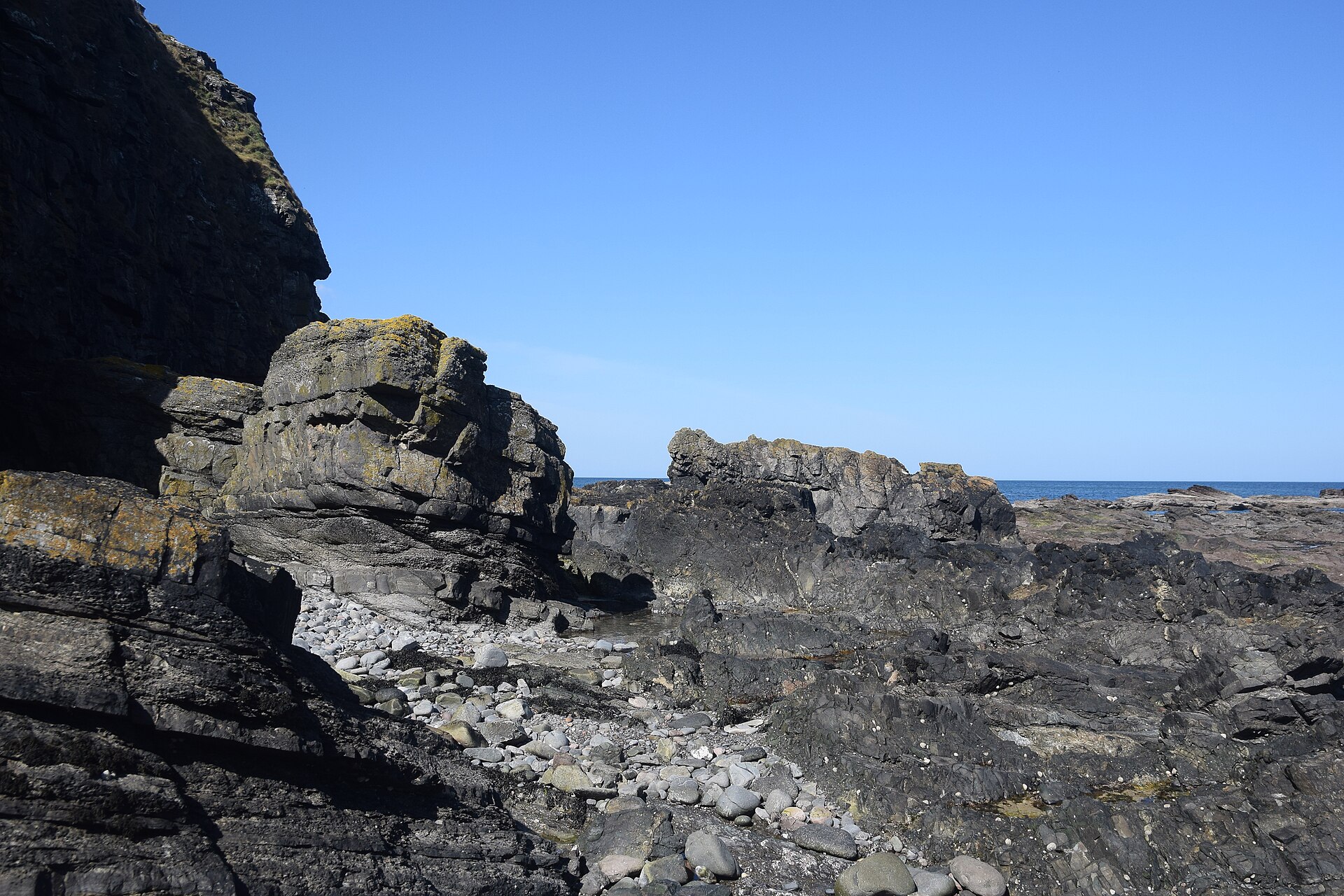 Dark rocks and reef along the shore at Aberdour Beach, the sea beyond.