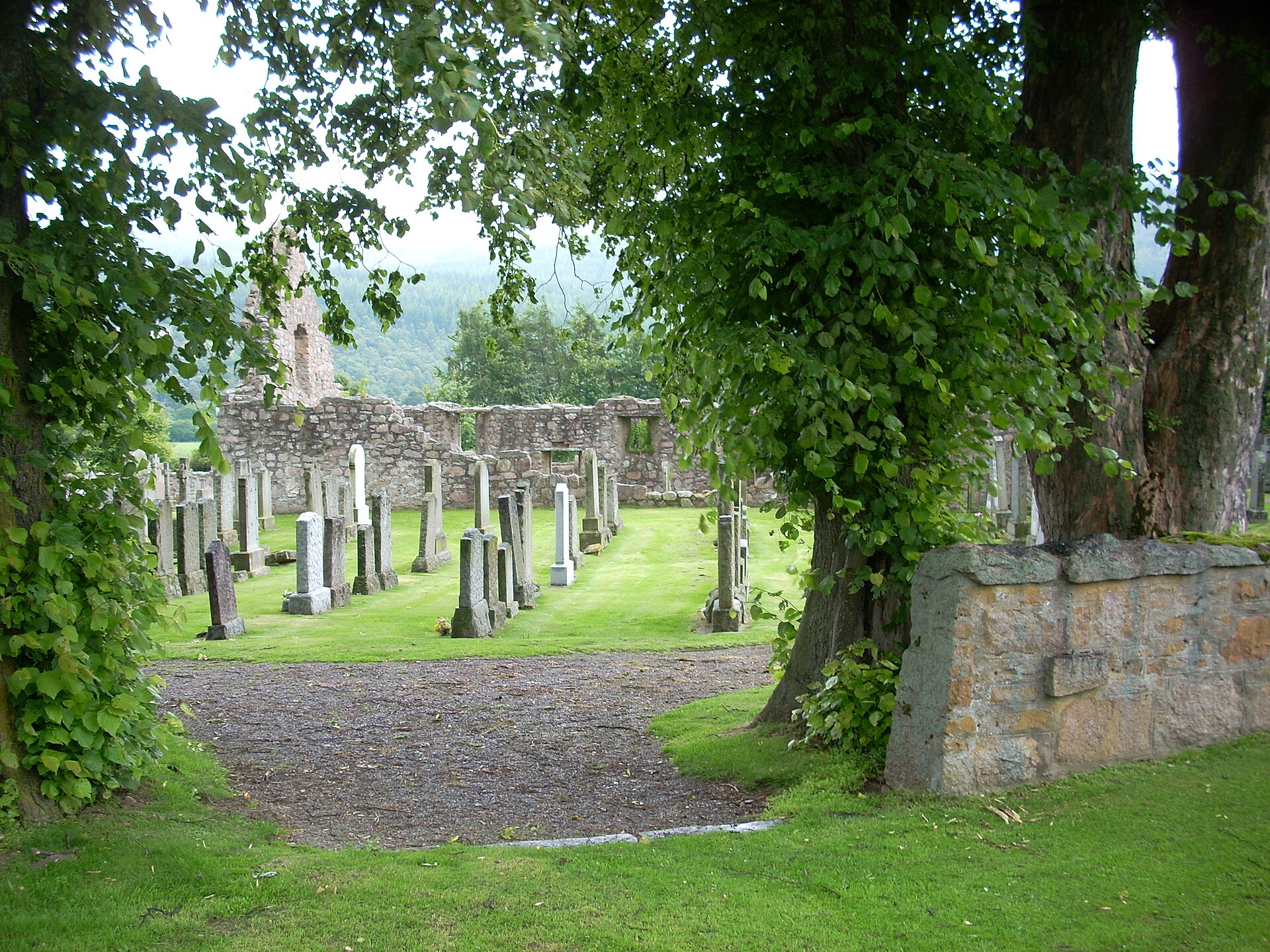 Old gravestones in the round kirkyard at Tullich among trees, the ruined kirk wall behind.