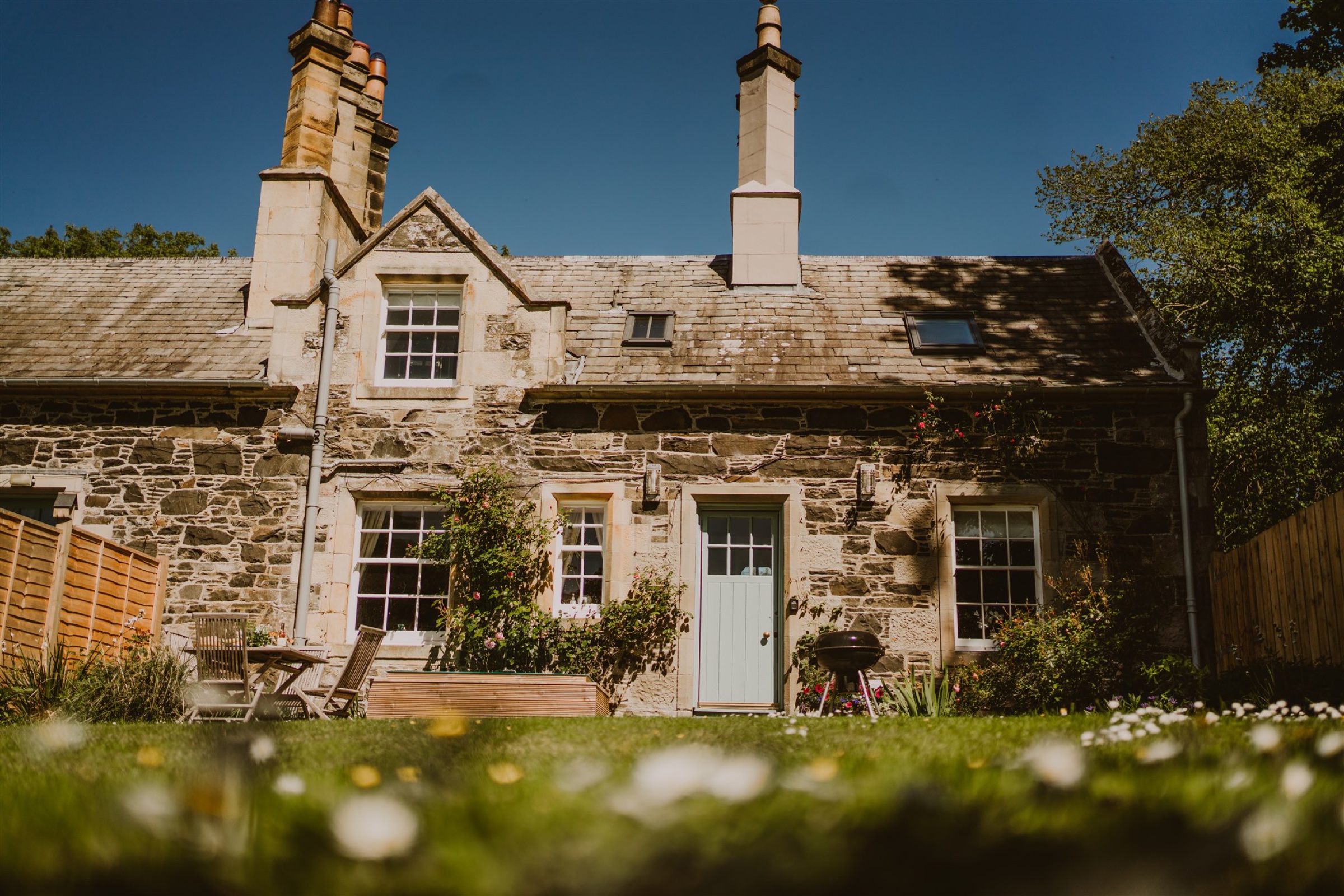 Fawn's Velvet Lodge in the sun, a stone cottage with a pale blue door, daisies on the lawn in front.