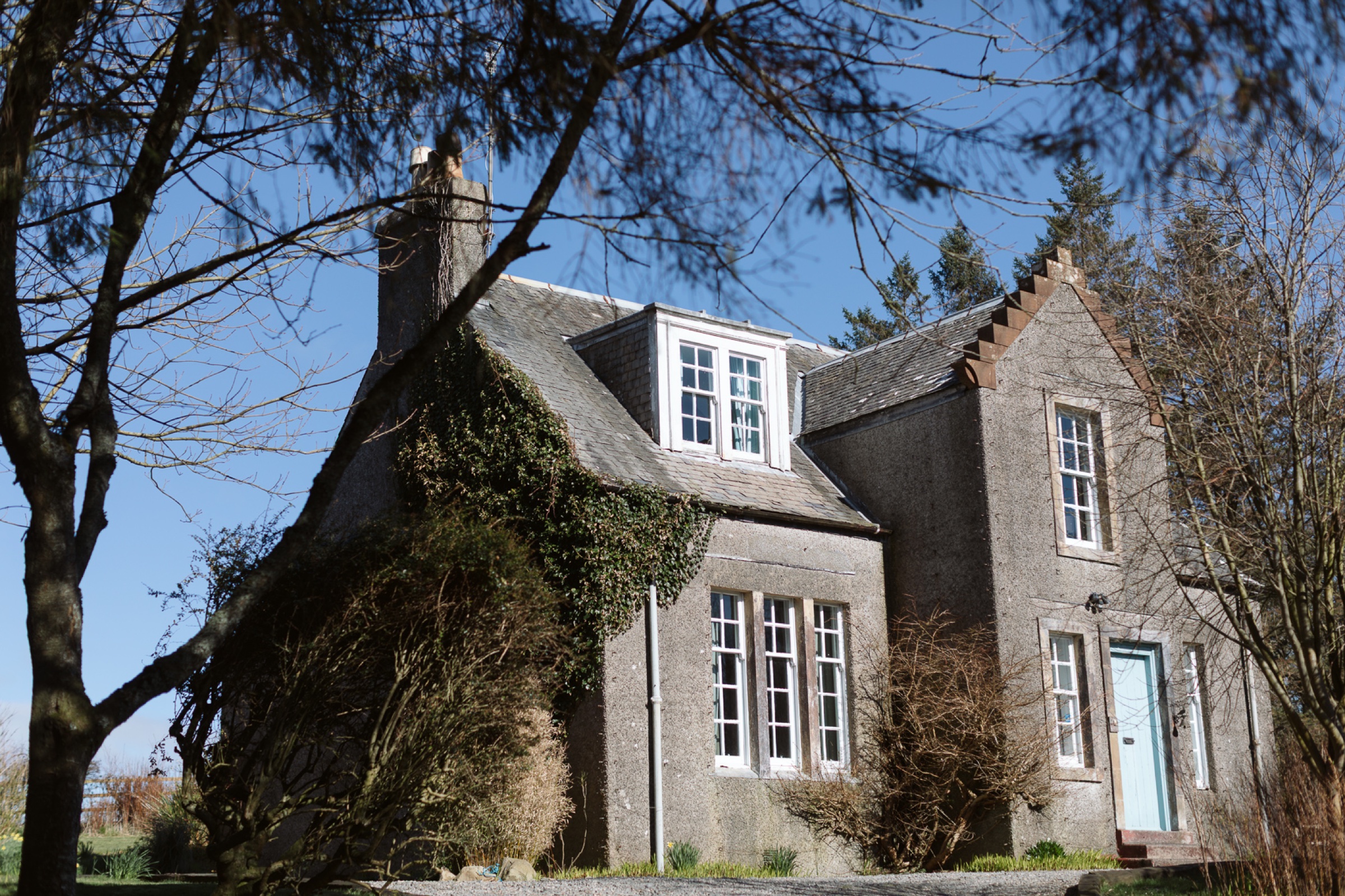 Fern Forest House, a grey stone house with ivy on its gable and a pale blue door, under a blue sky.