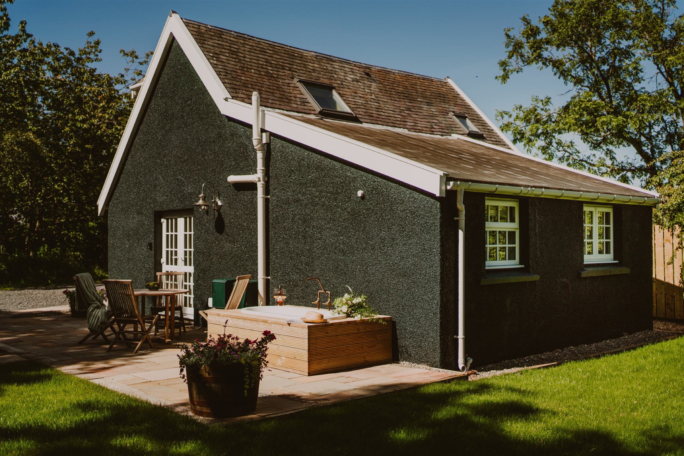 Harriet's Lodestar Lodge in the sun, dark walls and a white gable, the outdoor bath and a table on its terrace.