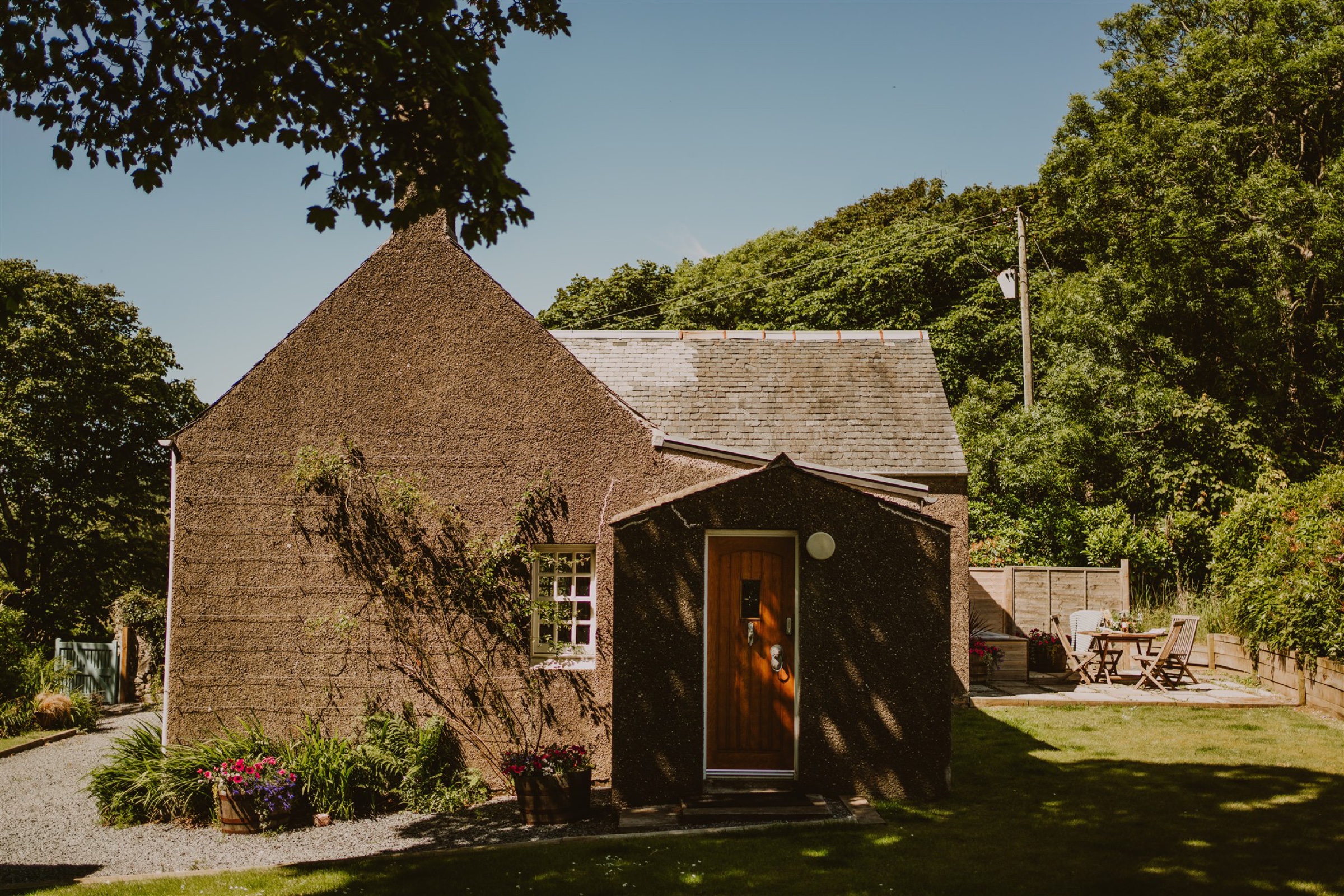 Rose Blair Lodge in the sun, a gabled cottage with a wooden door in a porch, trees behind and a table on the terrace.