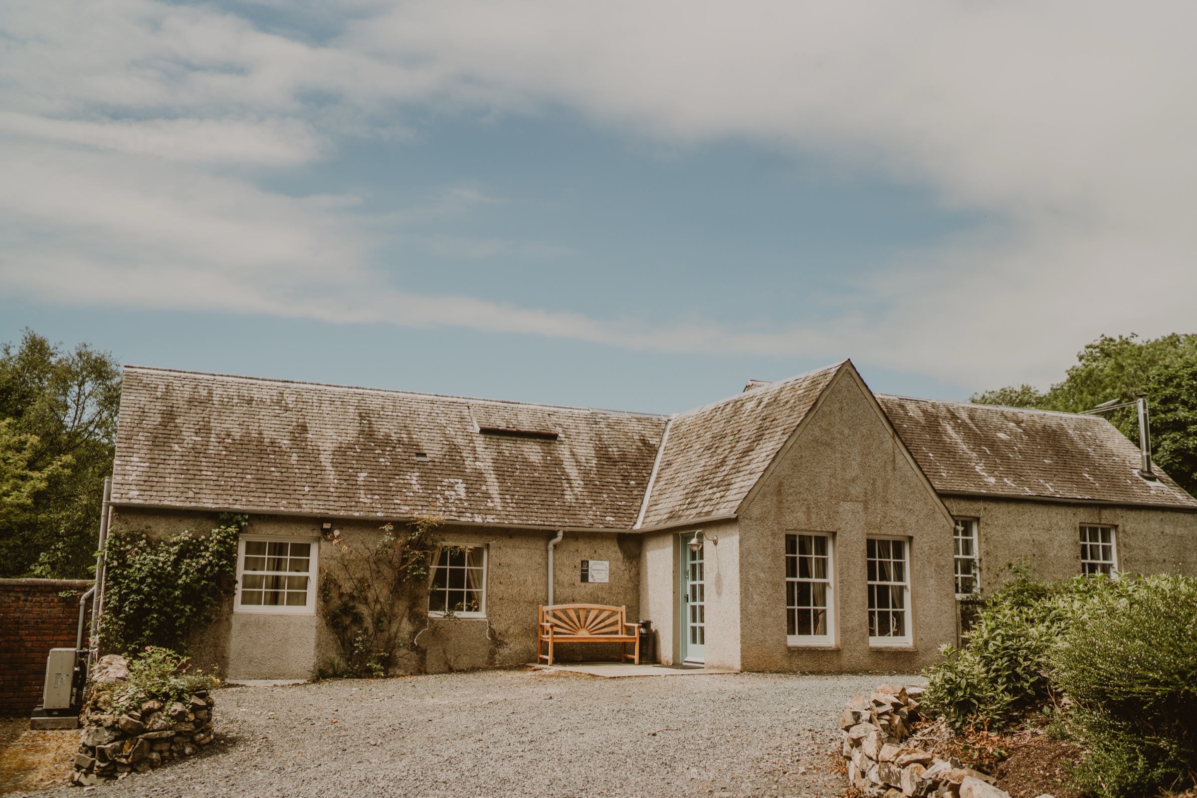 Symphonica Lodge, a long single-storey stone building with a bench by the door, on a gravel drive.