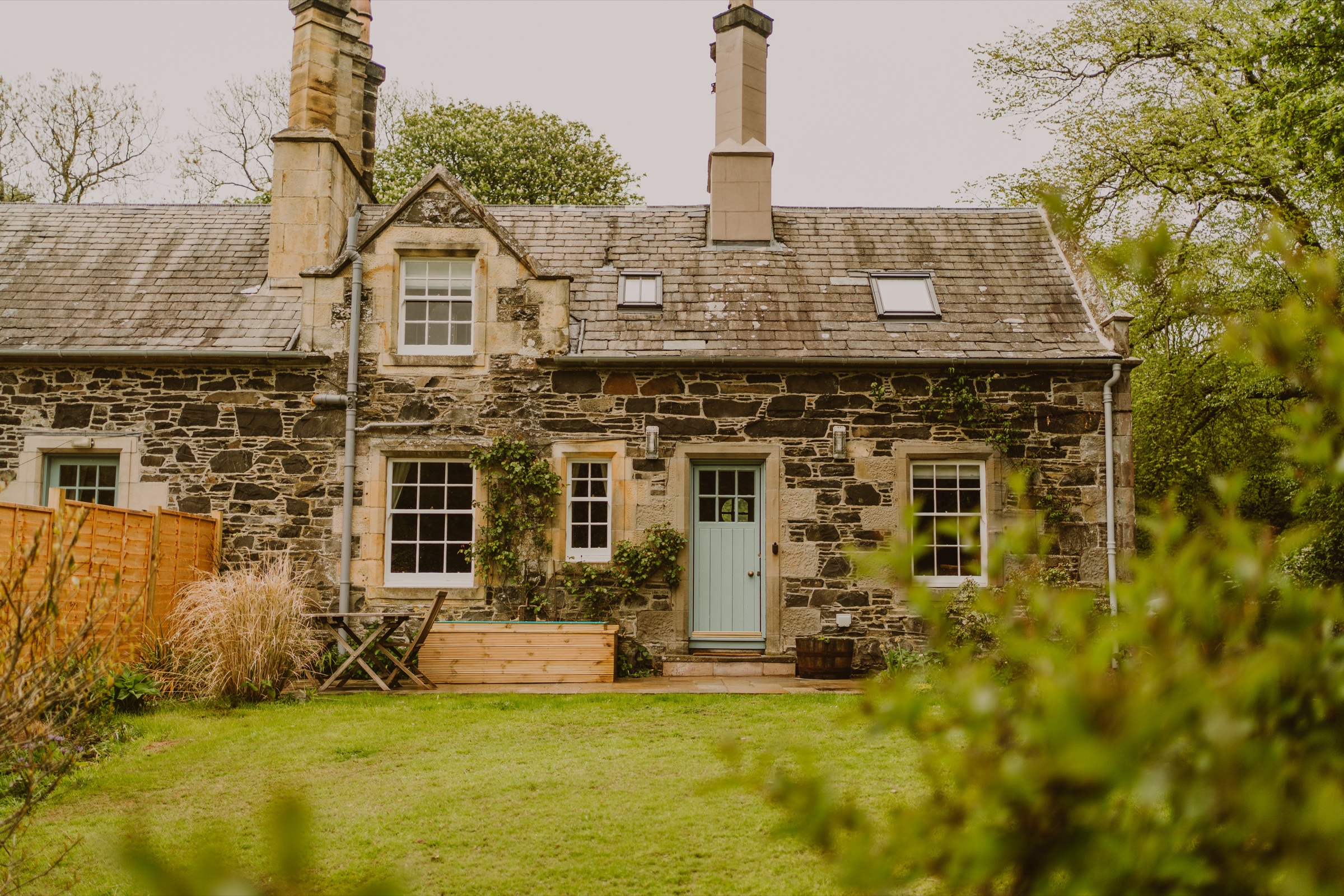 The front of Wren's Nest, a stone cottage with a pale blue door, a table and chairs and a timber planter on the lawn before it.