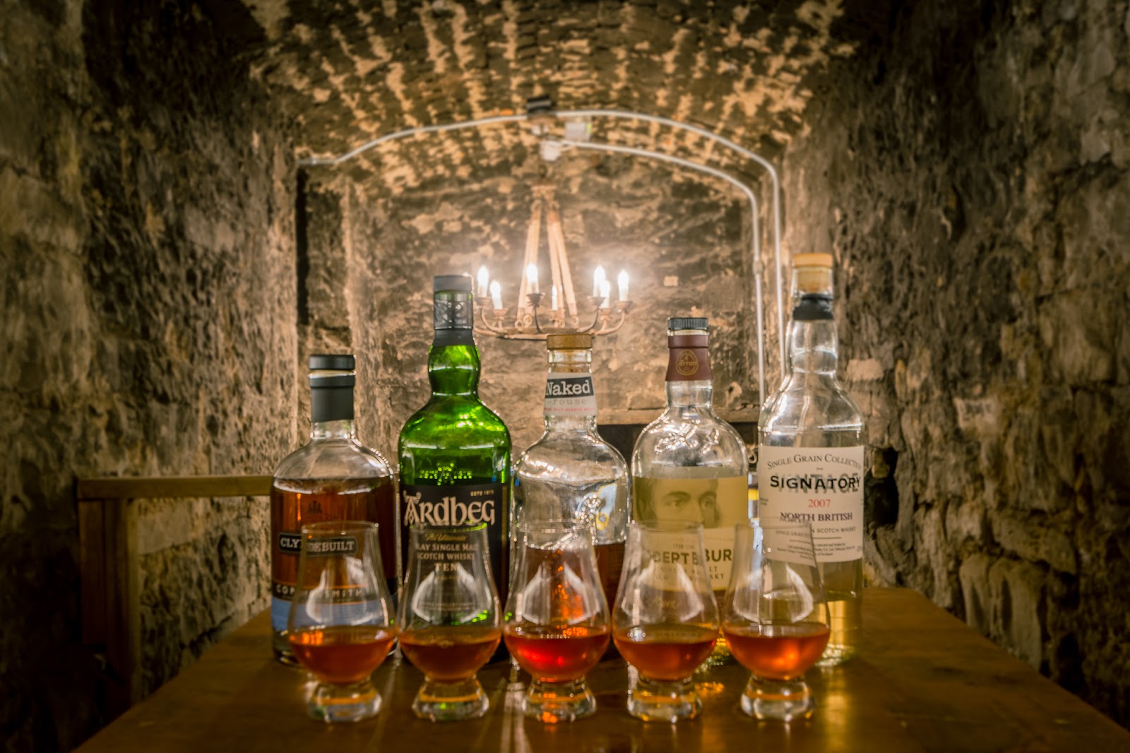 A flight of whiskies and their bottles on a table in a stone vault at the Lost Close, a chandelier behind.