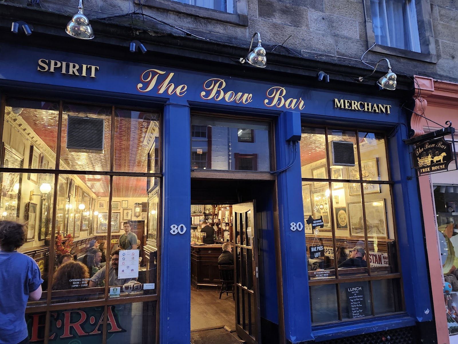 The blue front of The Bow Bar at 80 West Bow, its name in gold script between Spirit and Merchant, with the lit bar inside.
