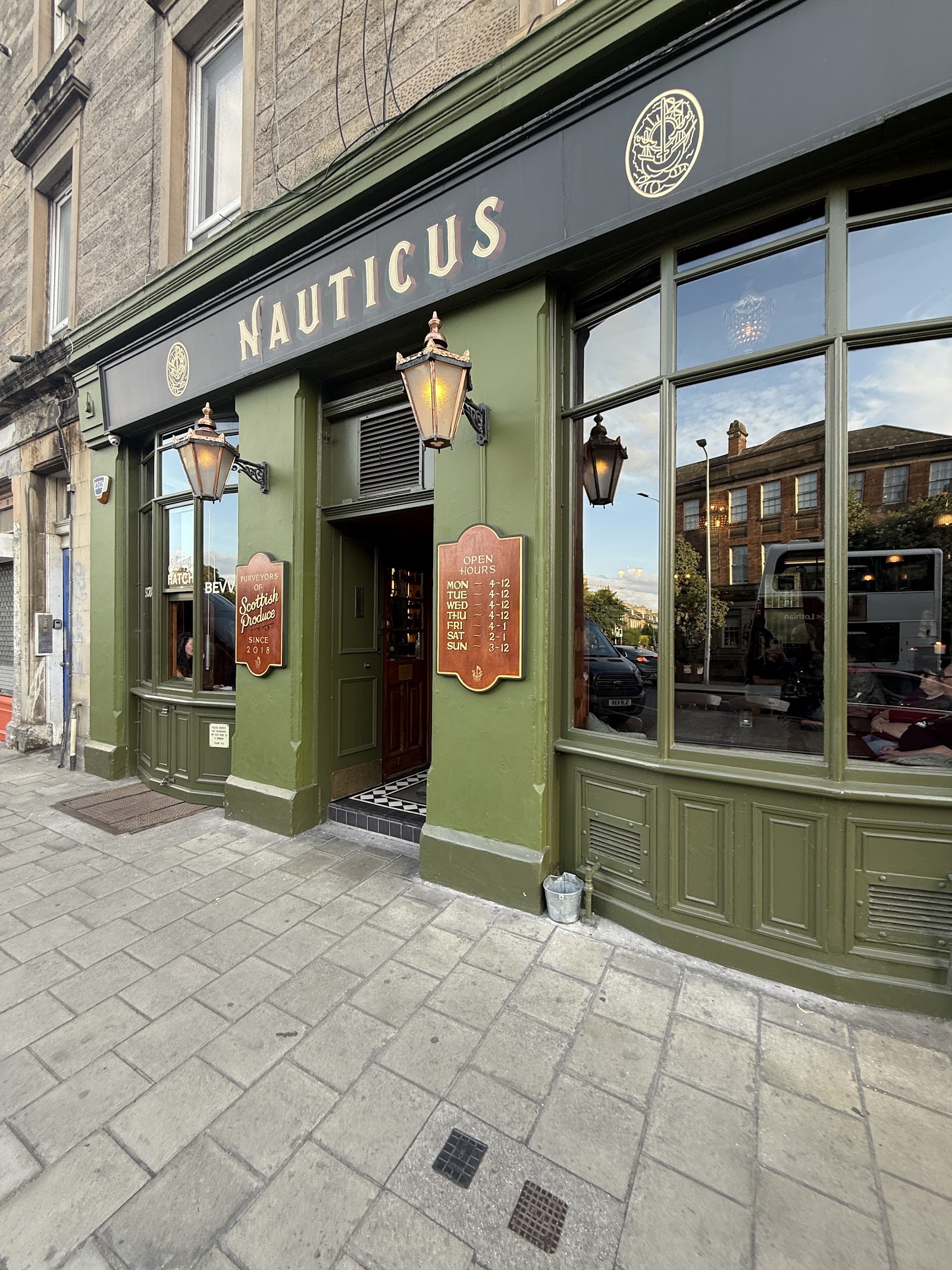 The green-painted front of Nauticus on Duke Street in Leith, with its name in gold above the door, brass lanterns and a sign reading Purveyors of Scottish Produce since 2018.
