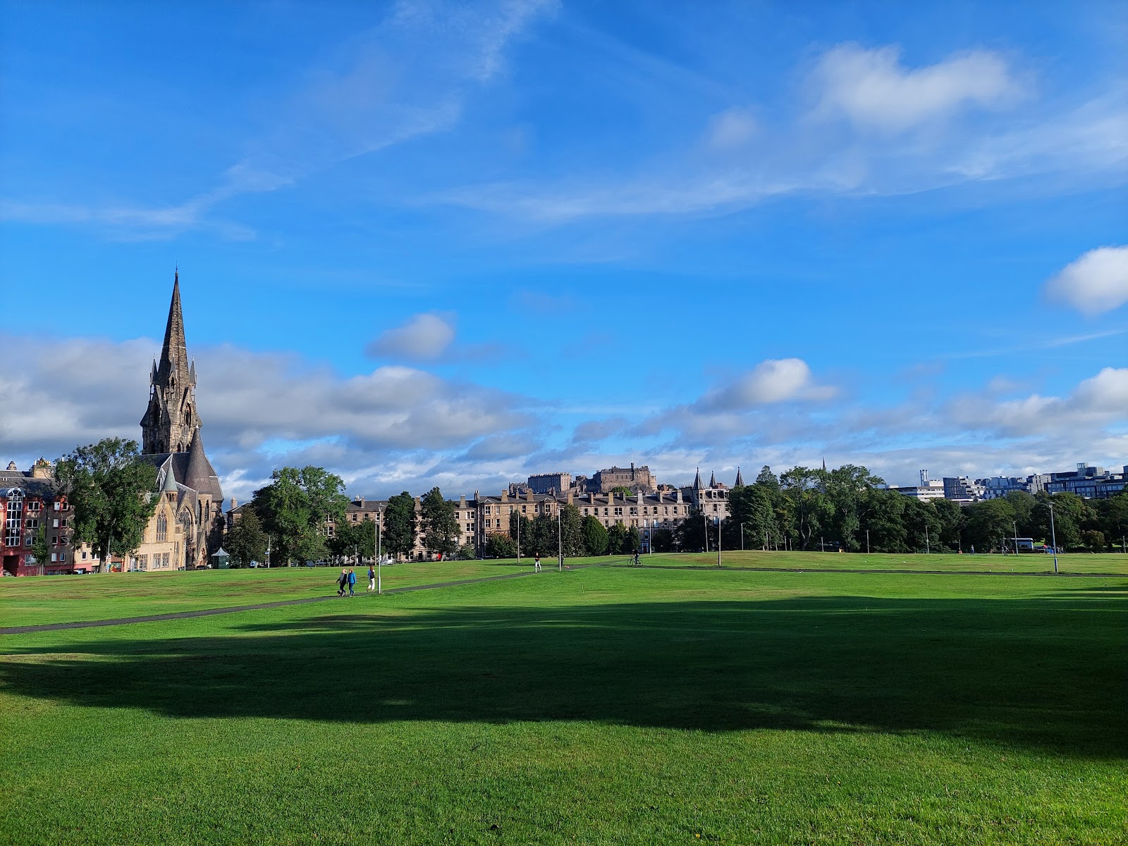 Open grass on Bruntsfield Links under a blue sky, a church spire and tenements beyond the trees.
