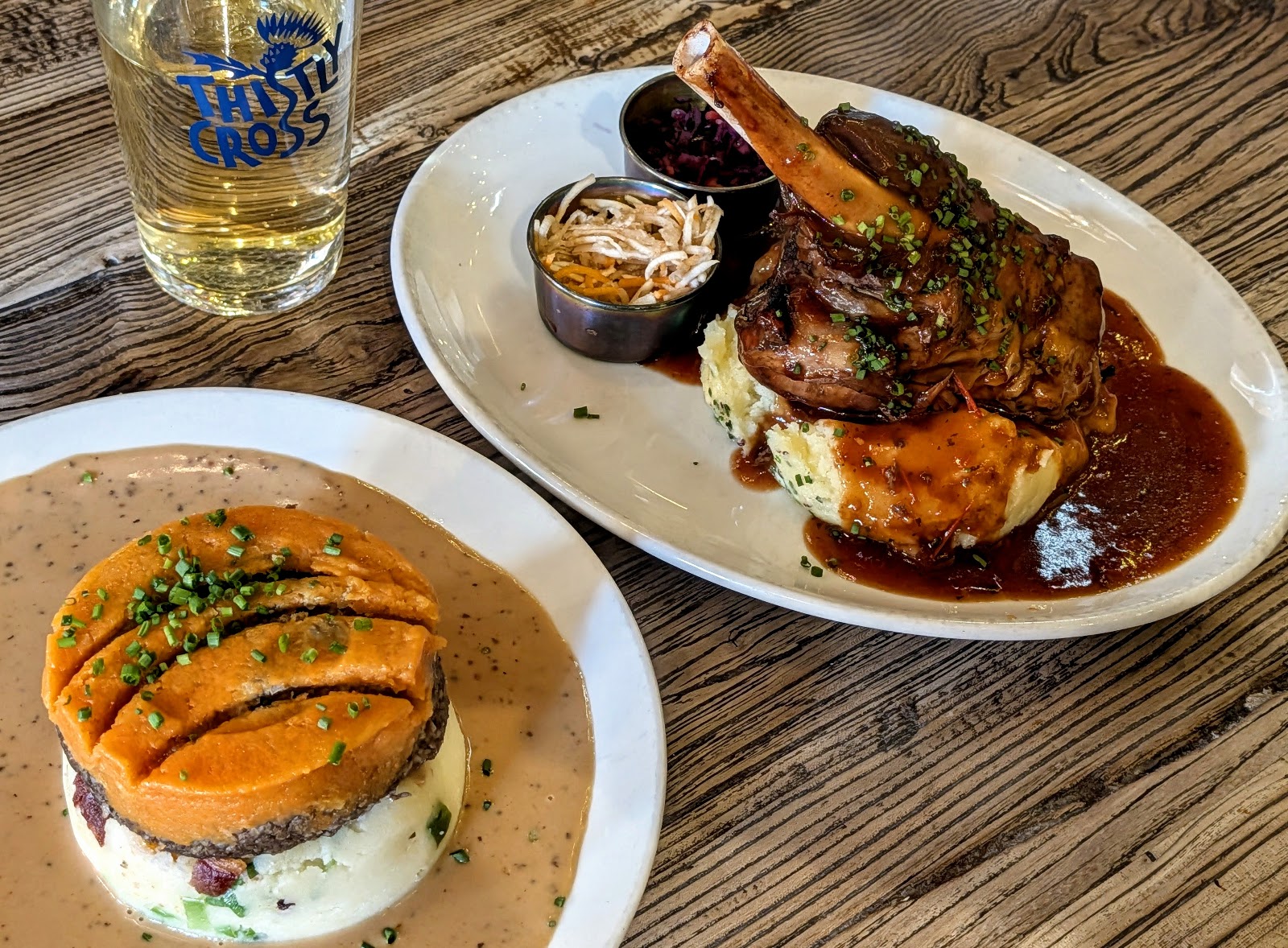 A lamb shank in gravy on mash beside a haggis stack topped with neeps, on a wooden table at Makars Mash Bar.