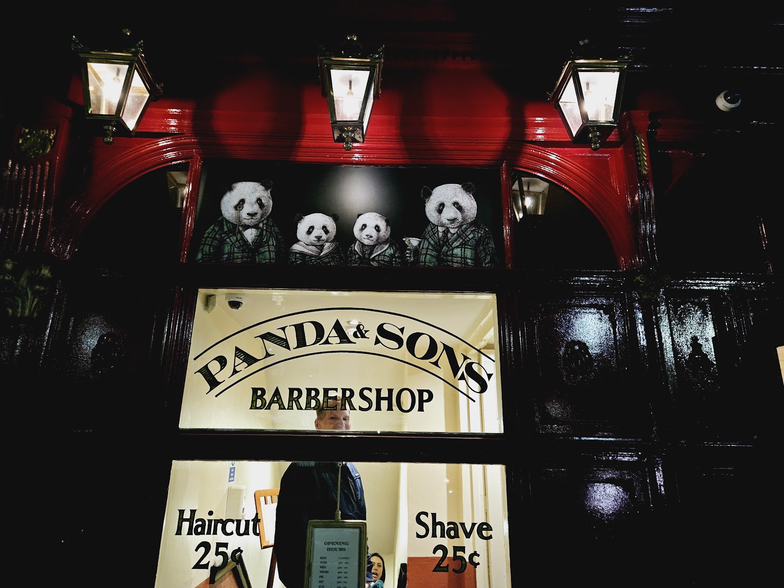 The red and black front of Panda & Sons on Queen Street at night, a barbershop window under a painting of four pandas in tartan.