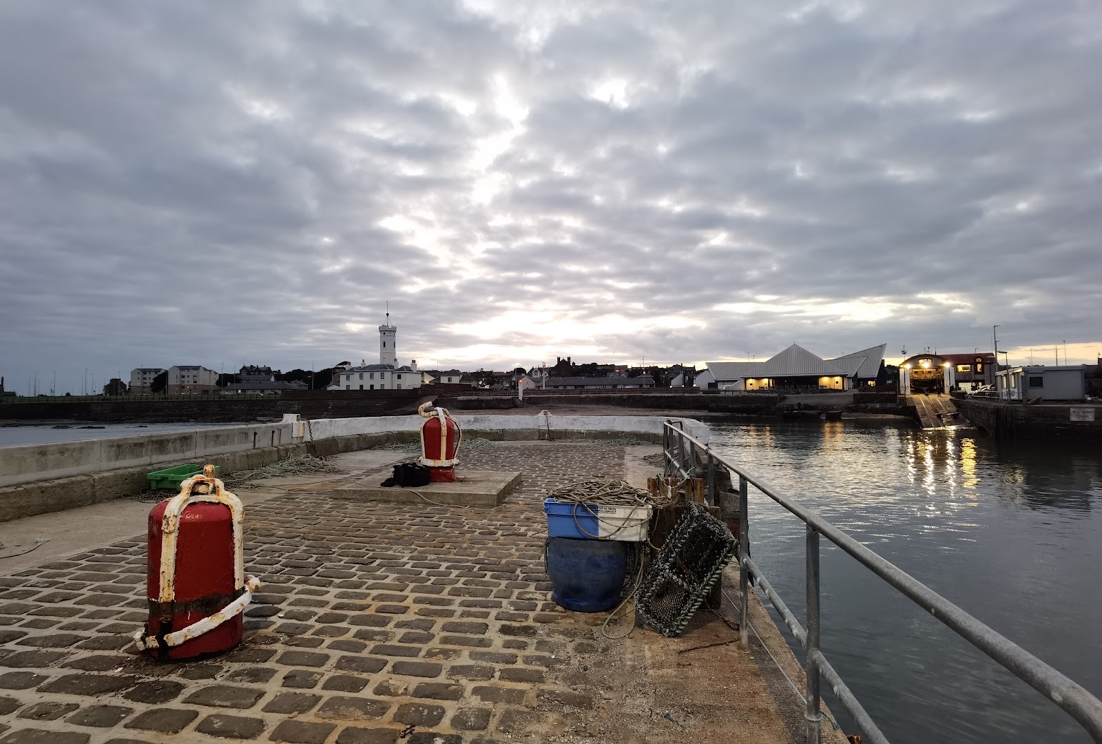 Red bollards and creels on a cobbled harbour pier, lighthouse in the distance