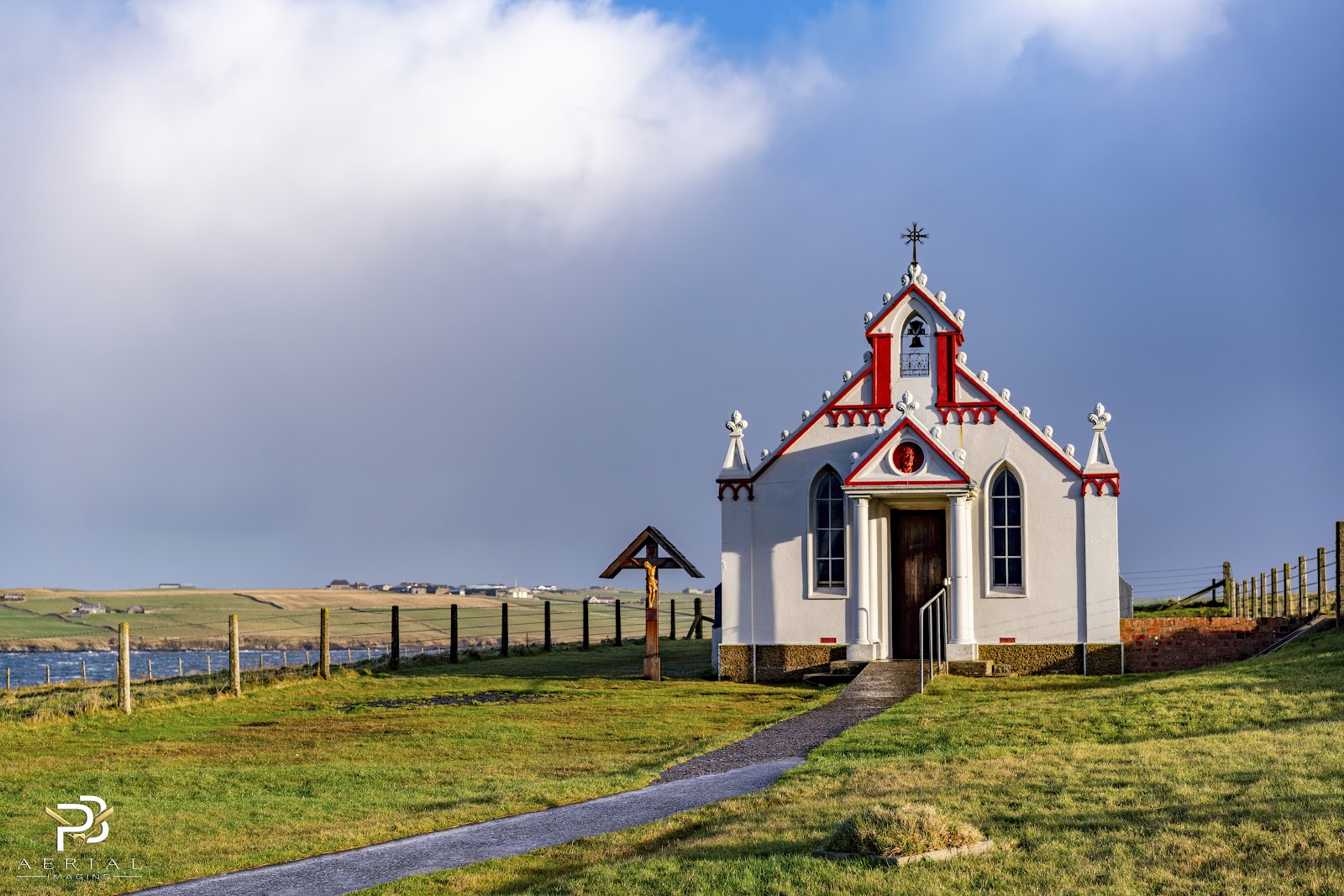 The white and red front of the Italian Chapel on Lamb Holm, a path leading up to it under a stormy sky.