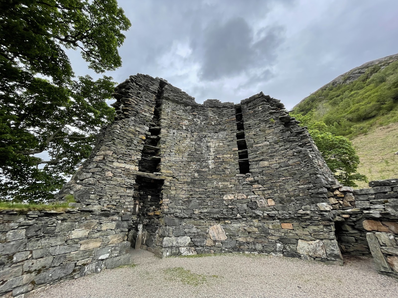 Drystone broch walls rise beside a wooded hillside under grey clouds