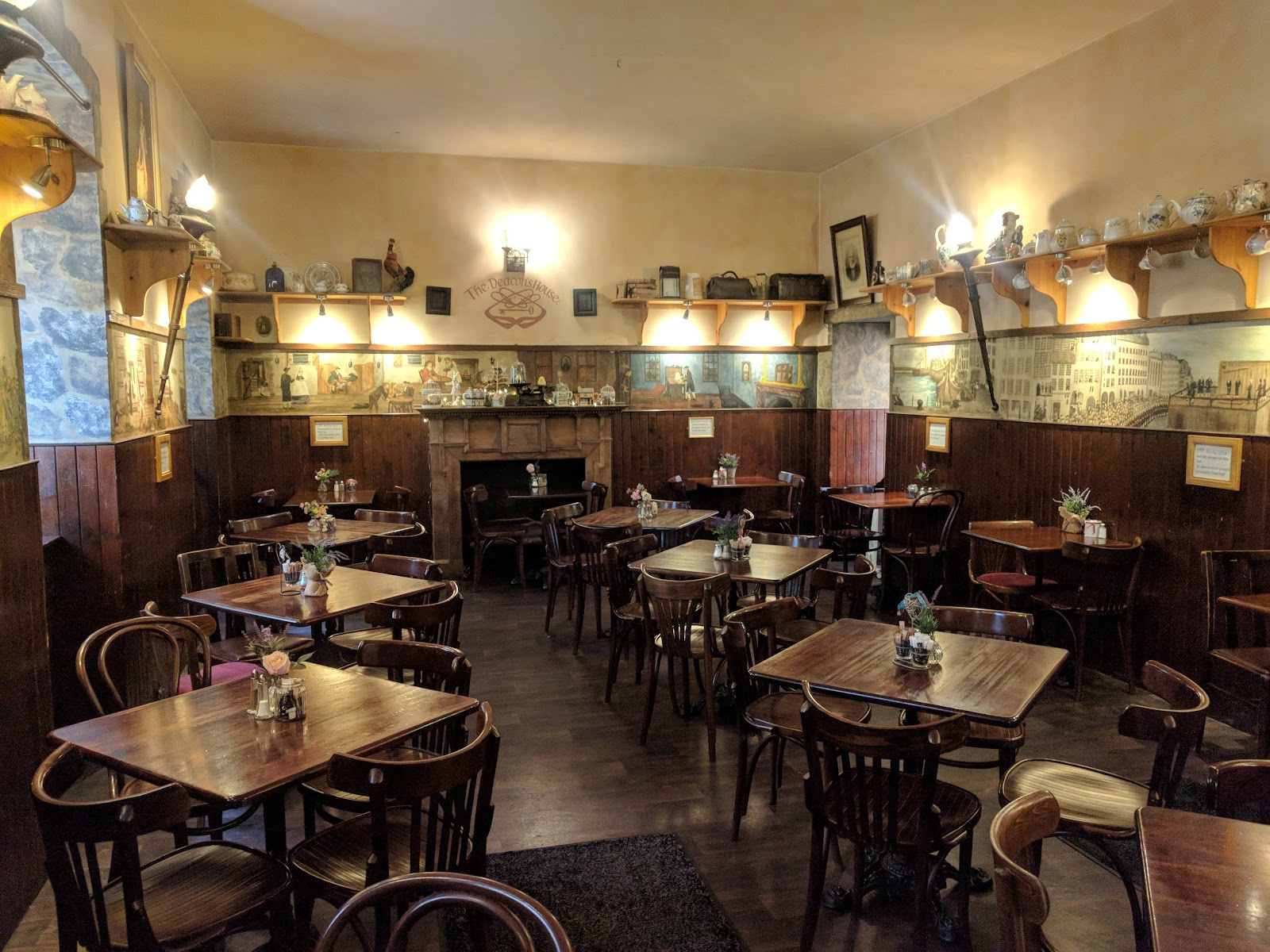 Empty tables and murals fill The Deacon's House tearoom, shelves lined with china above