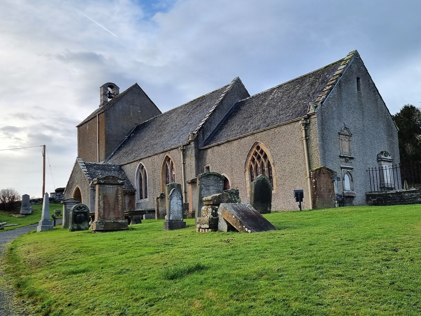 Stobo Kirk, a small grey stone church with a tower among gravestones on green grass under a blue sky.