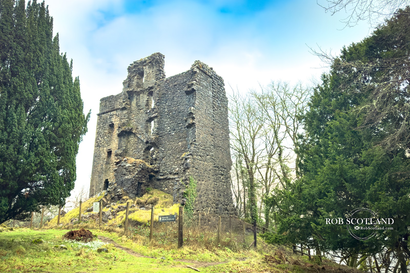 The tall roofless stone walls of Invergarry Castle among trees under a bright sky.