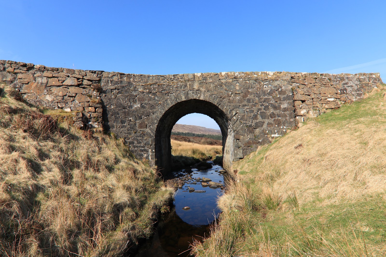 The Fairy Bridge on Skye: a low stone arch of dark rock carrying the road over a shallow burn, moorland and blue sky beyond.