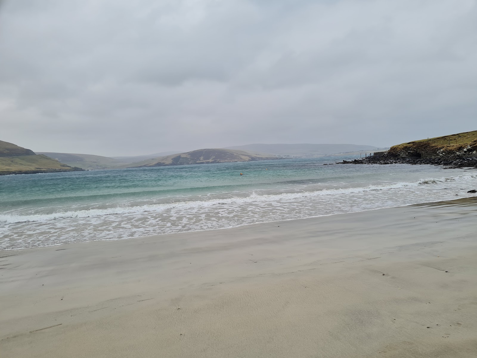 The pale sand and breaking waves of Levenwick bay, green headlands on either side.
