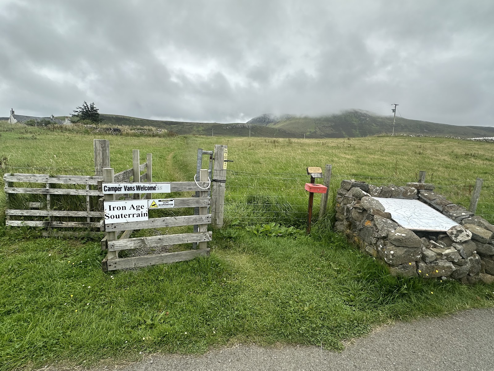 The Kilvaxter souterrain at Kilmuir: the stone-lined entrance open in the turf beside a fenced path, with the interpretation board behind it.
