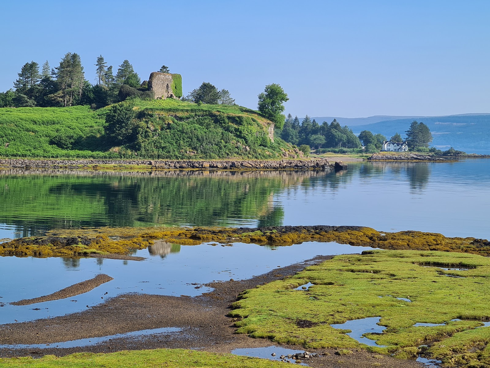 Aros Castle on its green knoll above a still bay at low tide, trees and a white house along the shore.