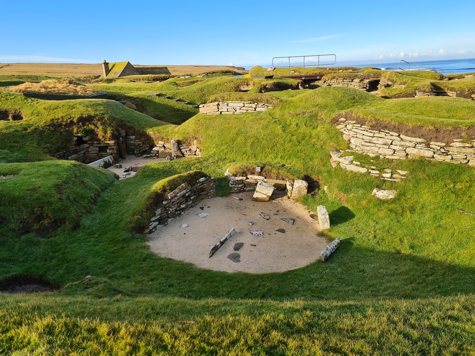 The grassy mounds and stone houses of Skara Brae in low evening sun, the sea behind.