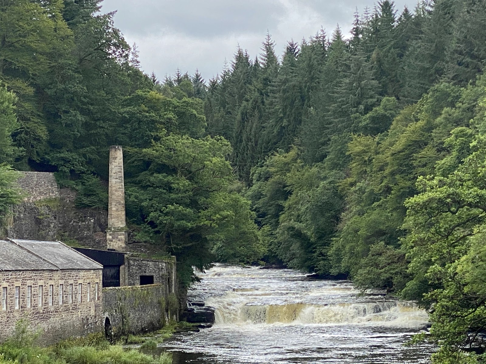 Dundaff Linn below the New Lanark mills, white water over a rock ledge between wooded banks, a tall chimney beside it.