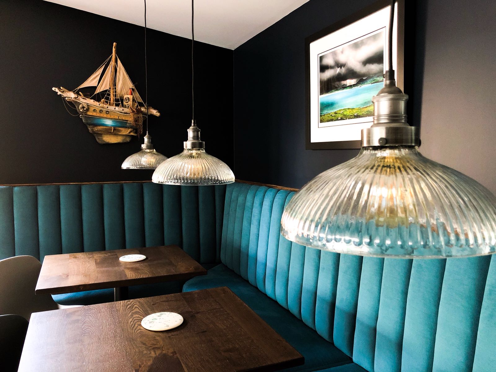 Teal banquettes and wooden tables at IV10 under glass pendant lamps and a model sailing ship.