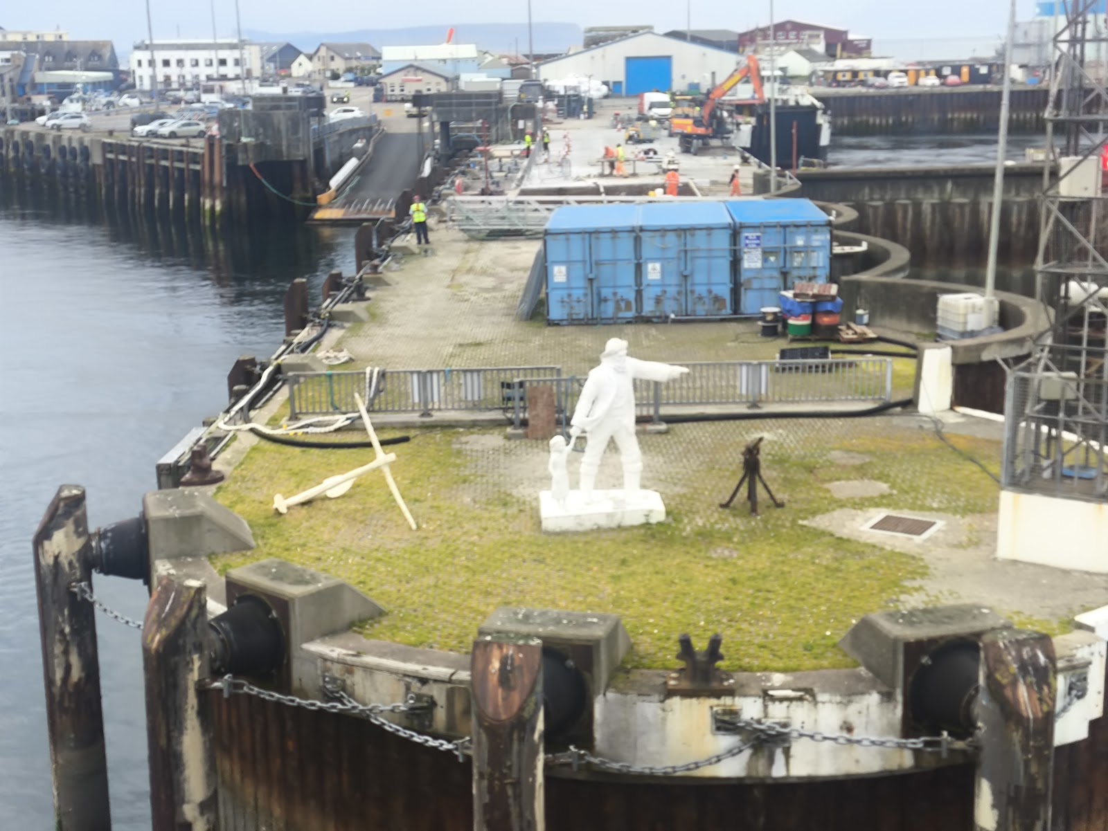 The white Fisherman and Child statue on a round grassy platform at the end of Mallaig's harbour wall, an anchor beside it and the harbour behind.
