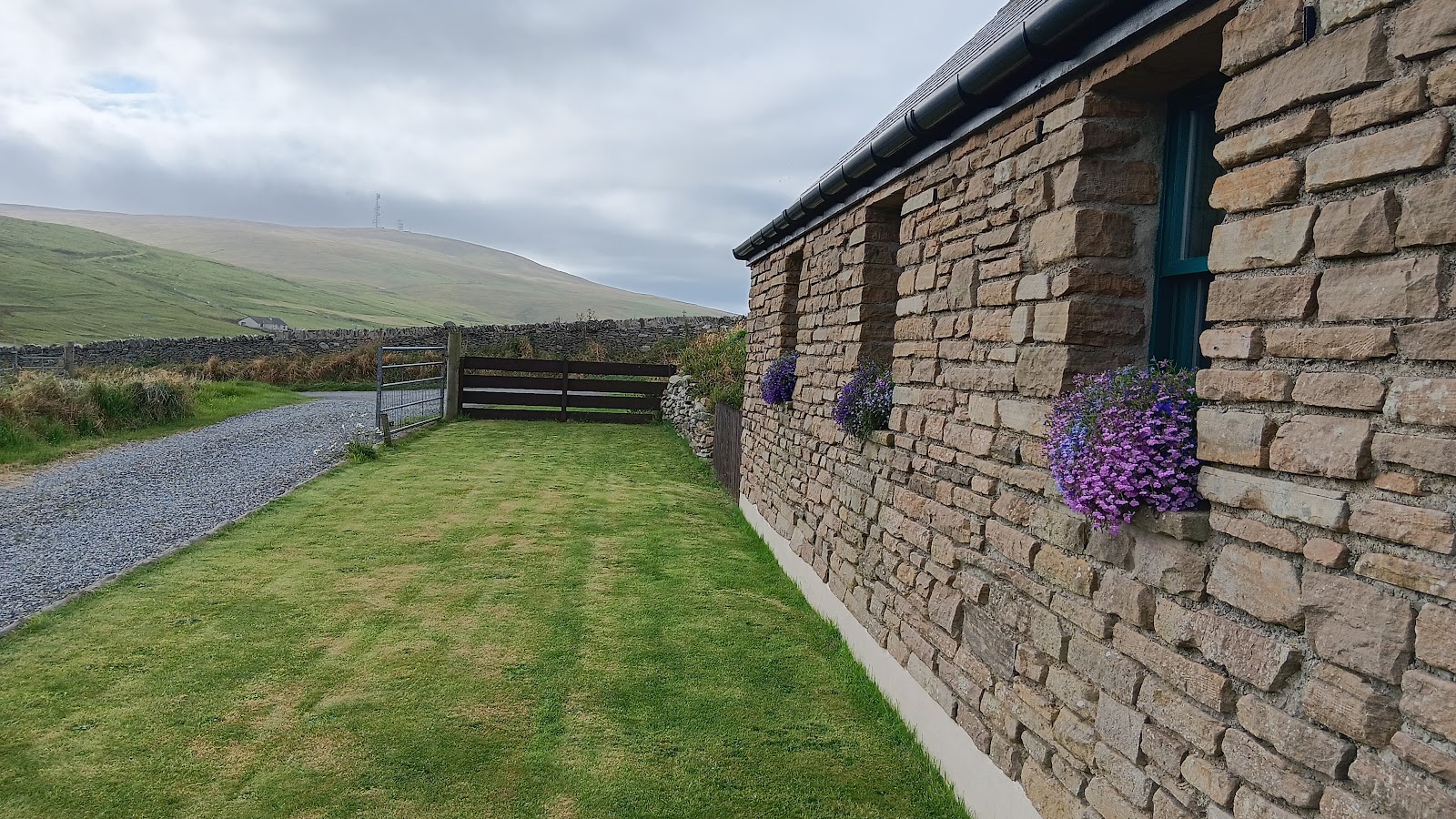 Purple flower baskets on a stone cottage wall overlooking hills and a gravel track