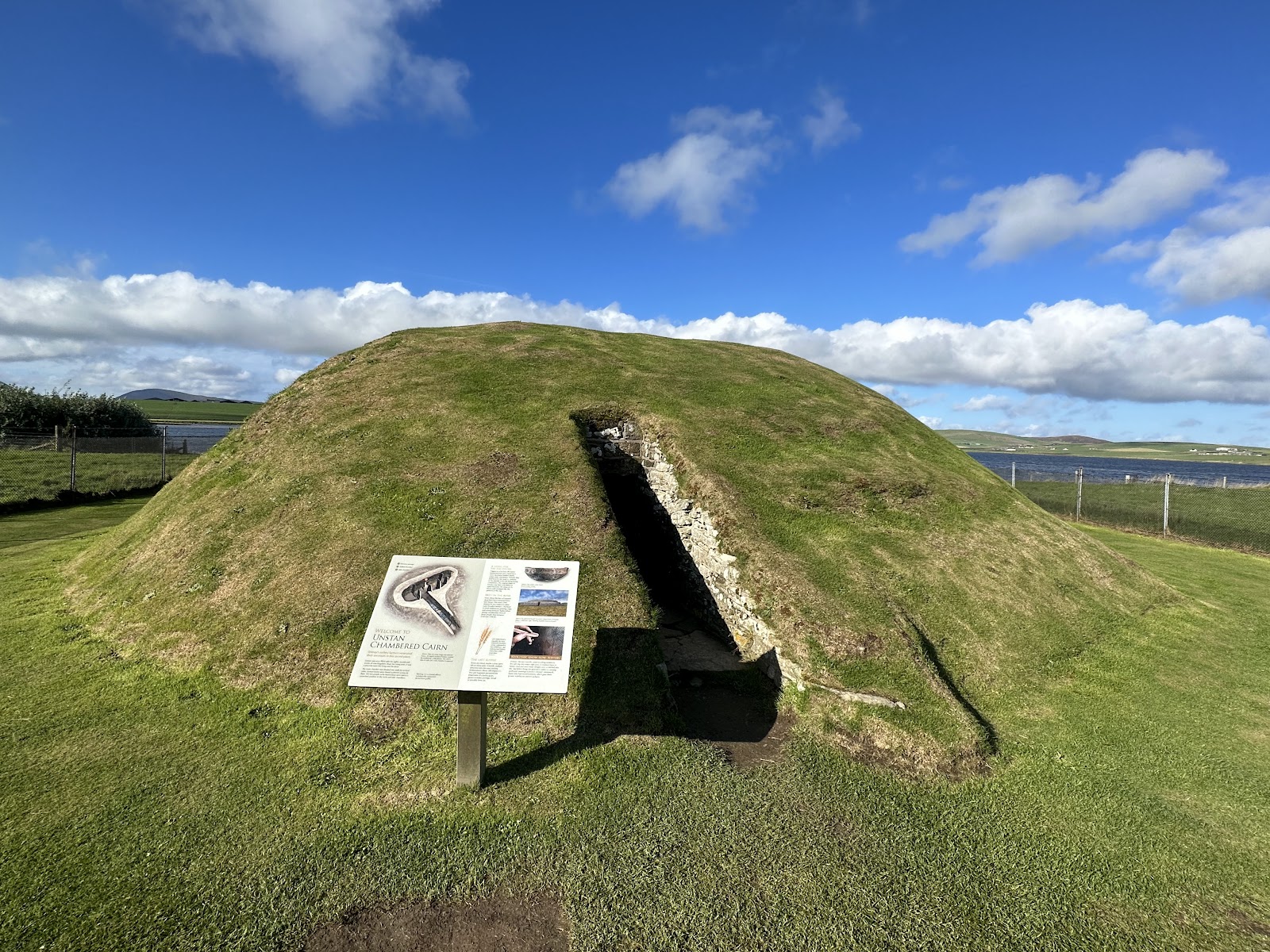 Unstan Chambered Cairn, a grassy mound with its low entrance and an information board, the loch beyond.