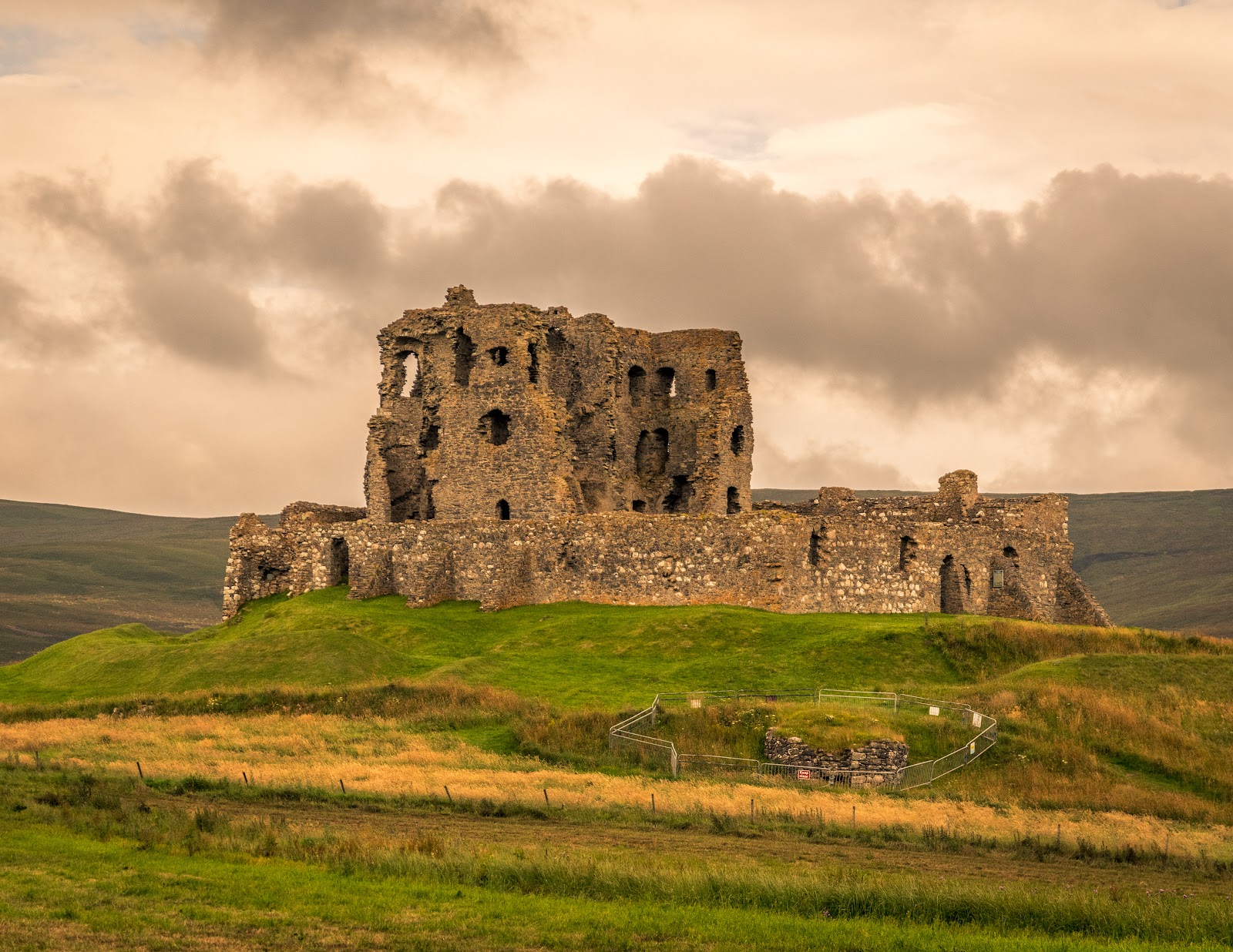 Ruined stone castle atop a grassy mound beneath a cloudy sky