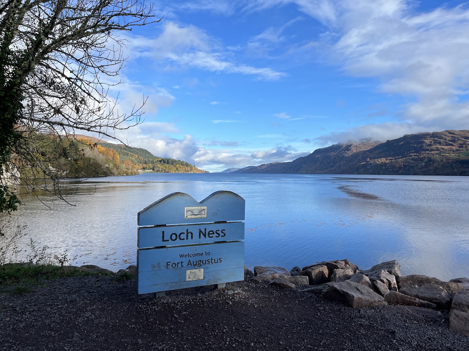 A sign reading Loch Ness, Welcome to Fort Augustus on the stony shore, the loch running north between wooded hills.