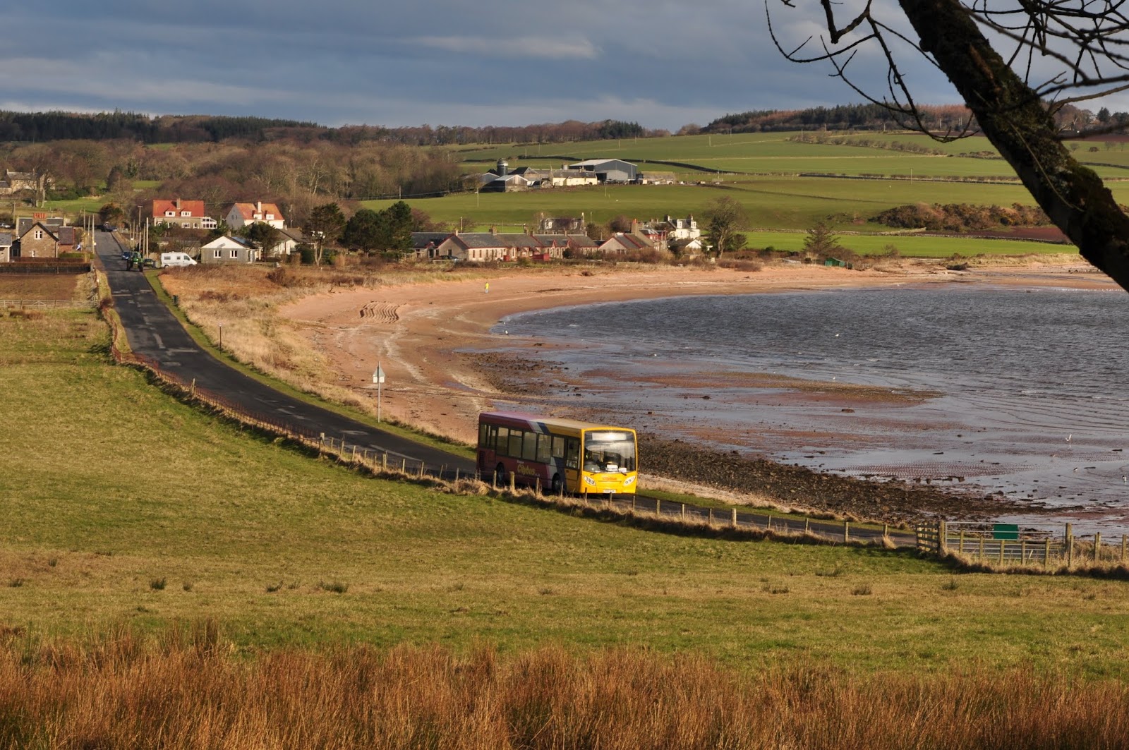 A bus on the shore road round the wide sandy bay, the village beyond.