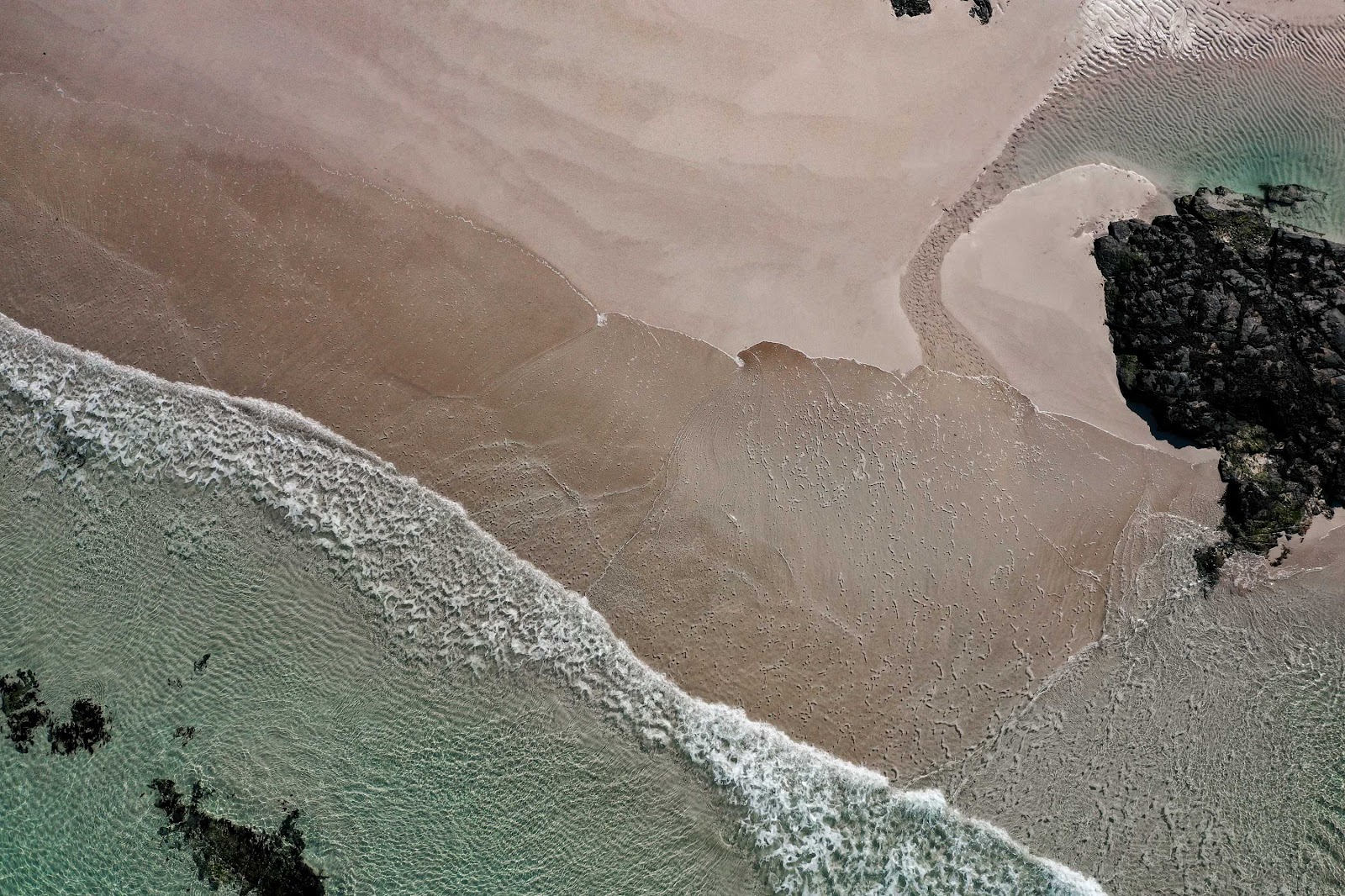 Aerial view of waves breaking on sand beside dark rocky outcrops