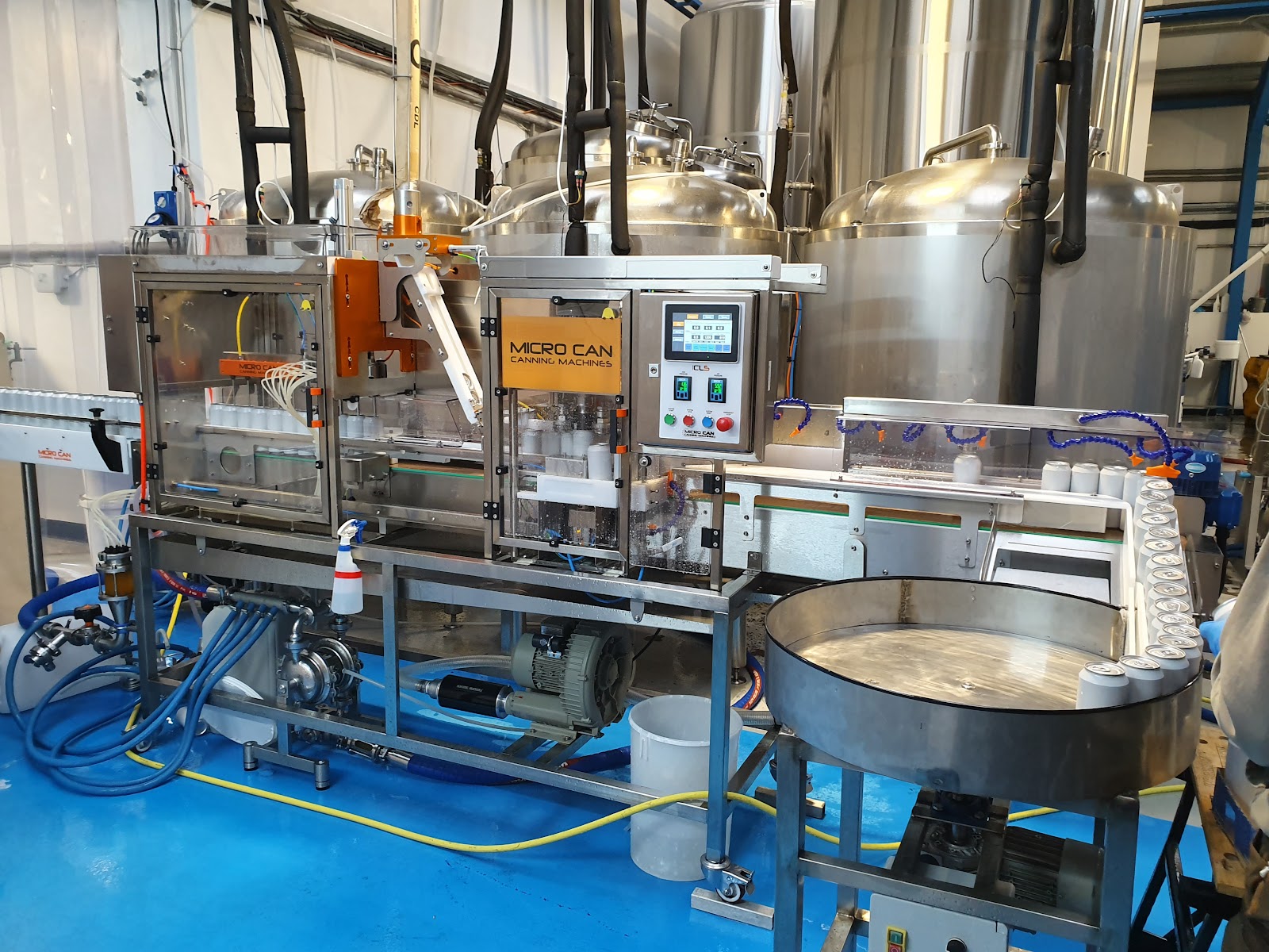 A Micro Can canning machine fills tins beside steel brewery fermentation tanks