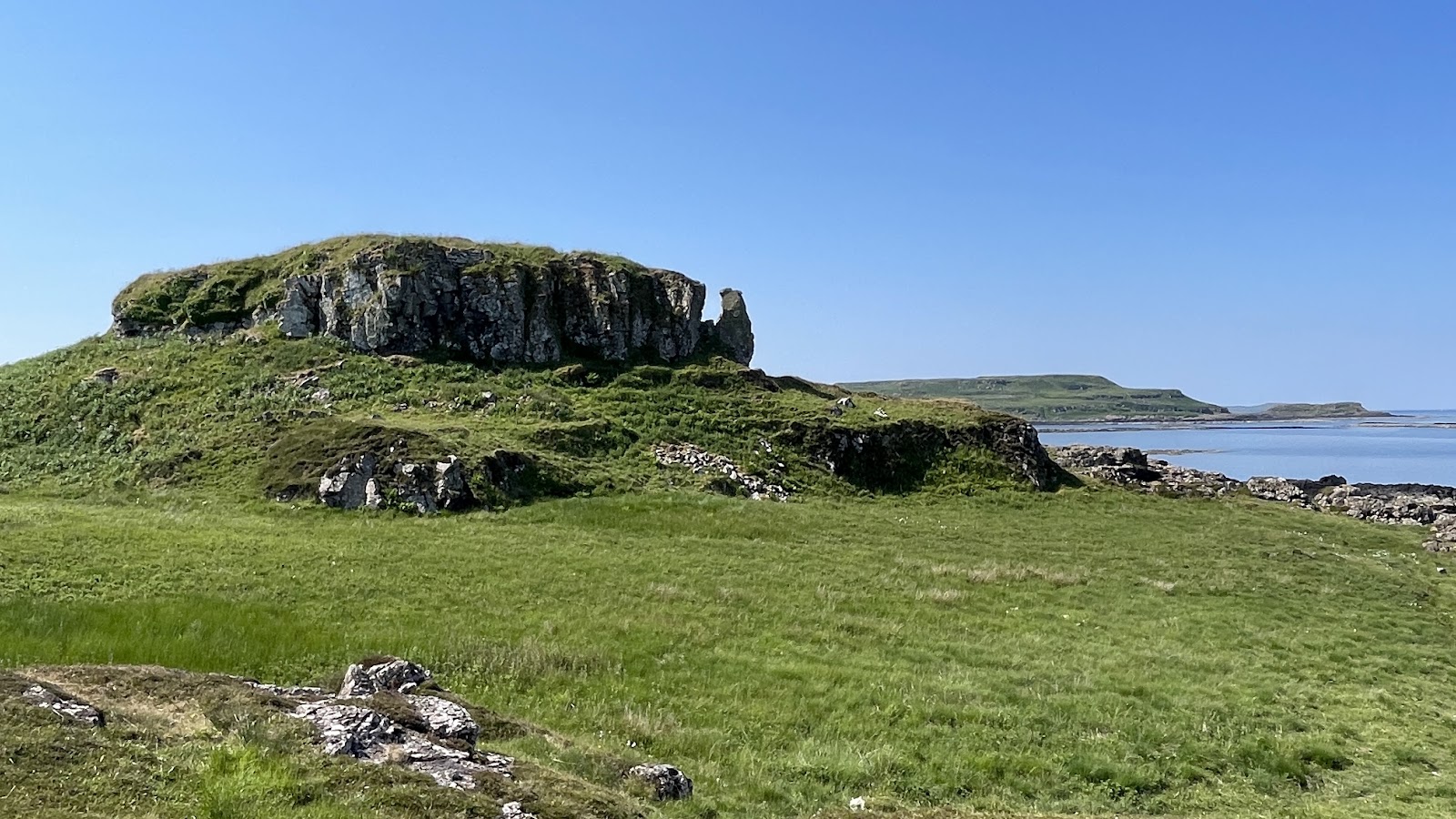 Dùn Ara's crag across a green field, the sea and hills beyond under a blue sky.