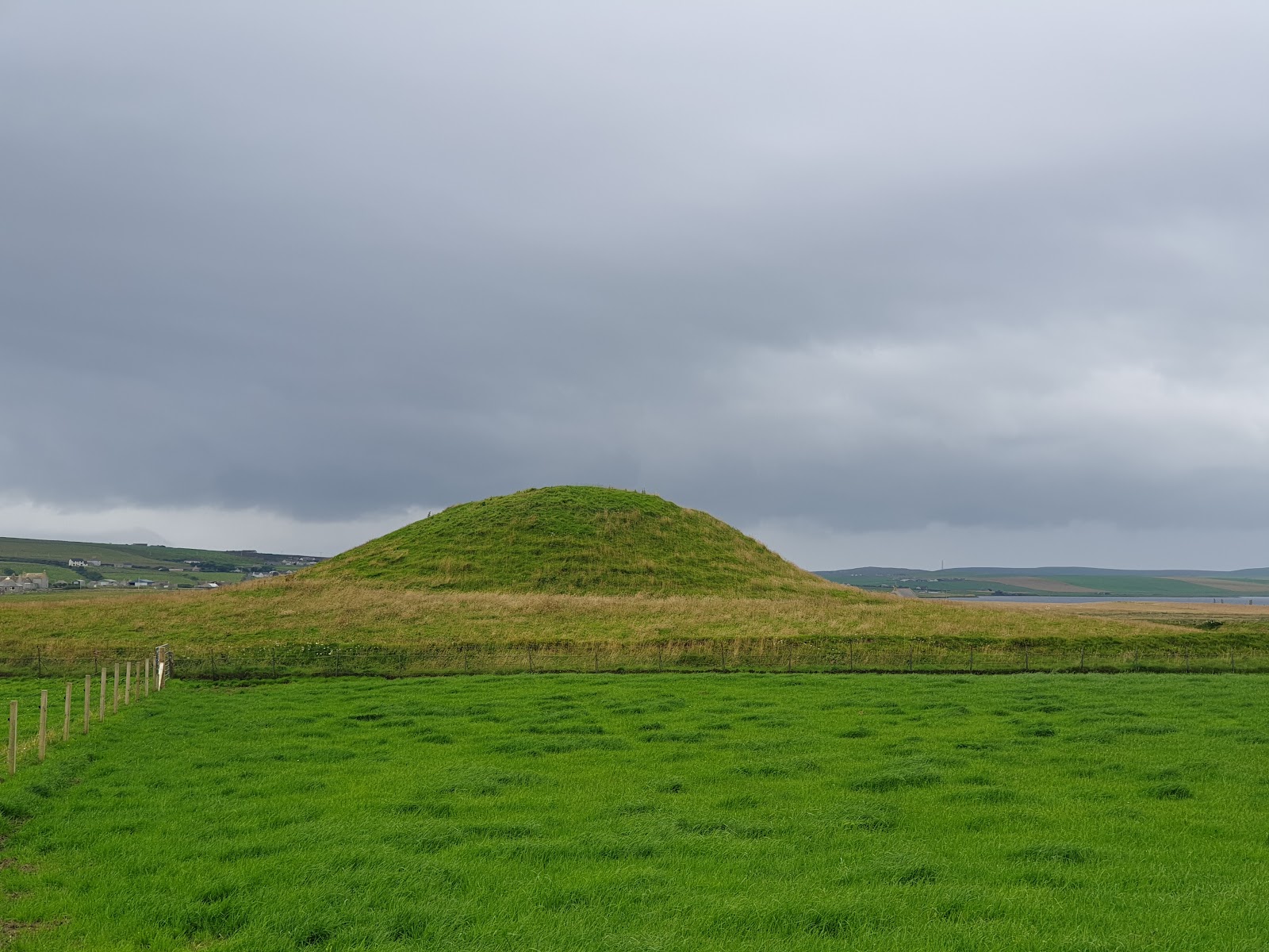 Maeshowe, a great grassy mound in green fields under a grey sky.