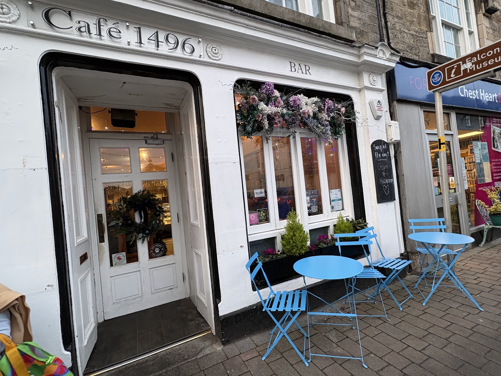 Cafe 1496 on Forres High Street, a white shopfront with gold lettering, a wreath on the door and blue tables outside.