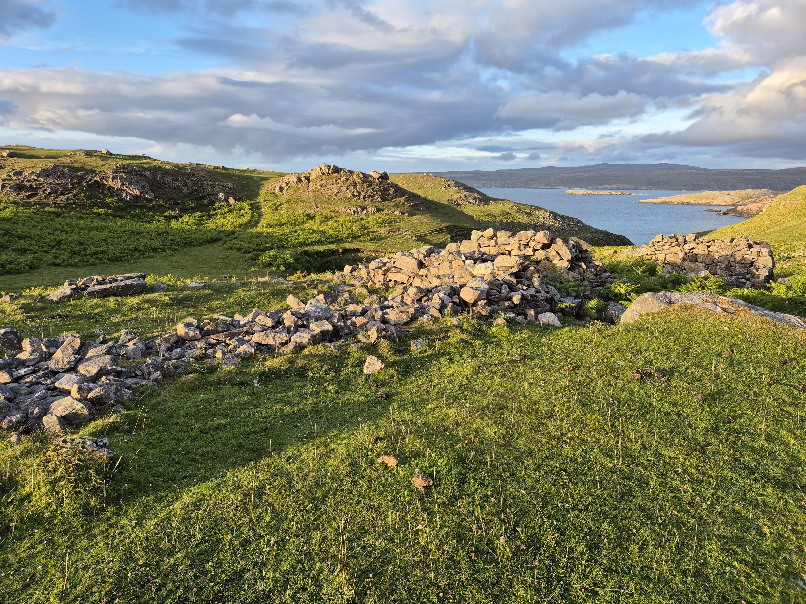 Tumbled stone walls of Ceannabeinne township on a green slope in evening light, the sea beyond.