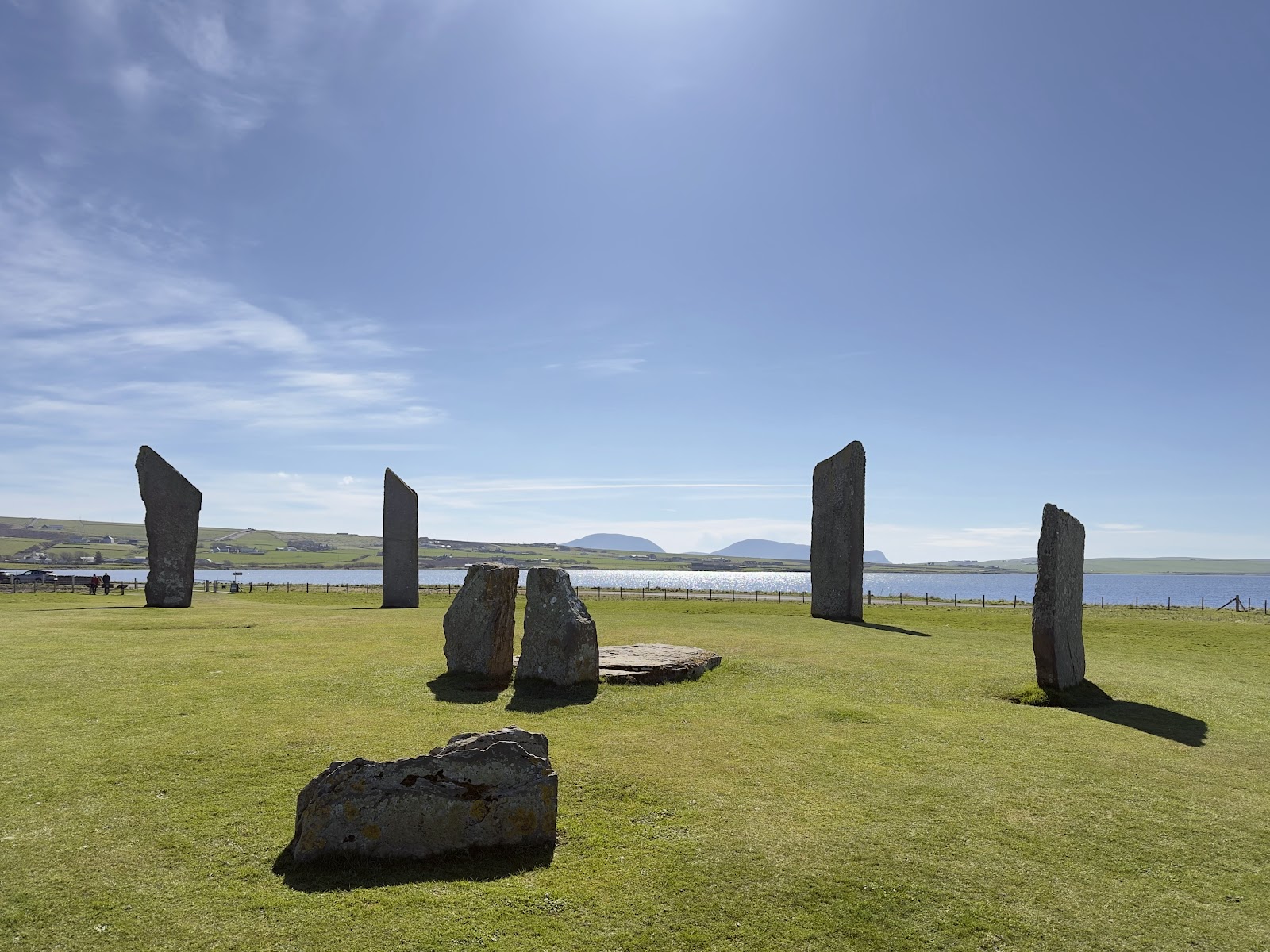The tall, thin Standing Stones of Stenness on green grass, the loch and the hills of Hoy behind.
